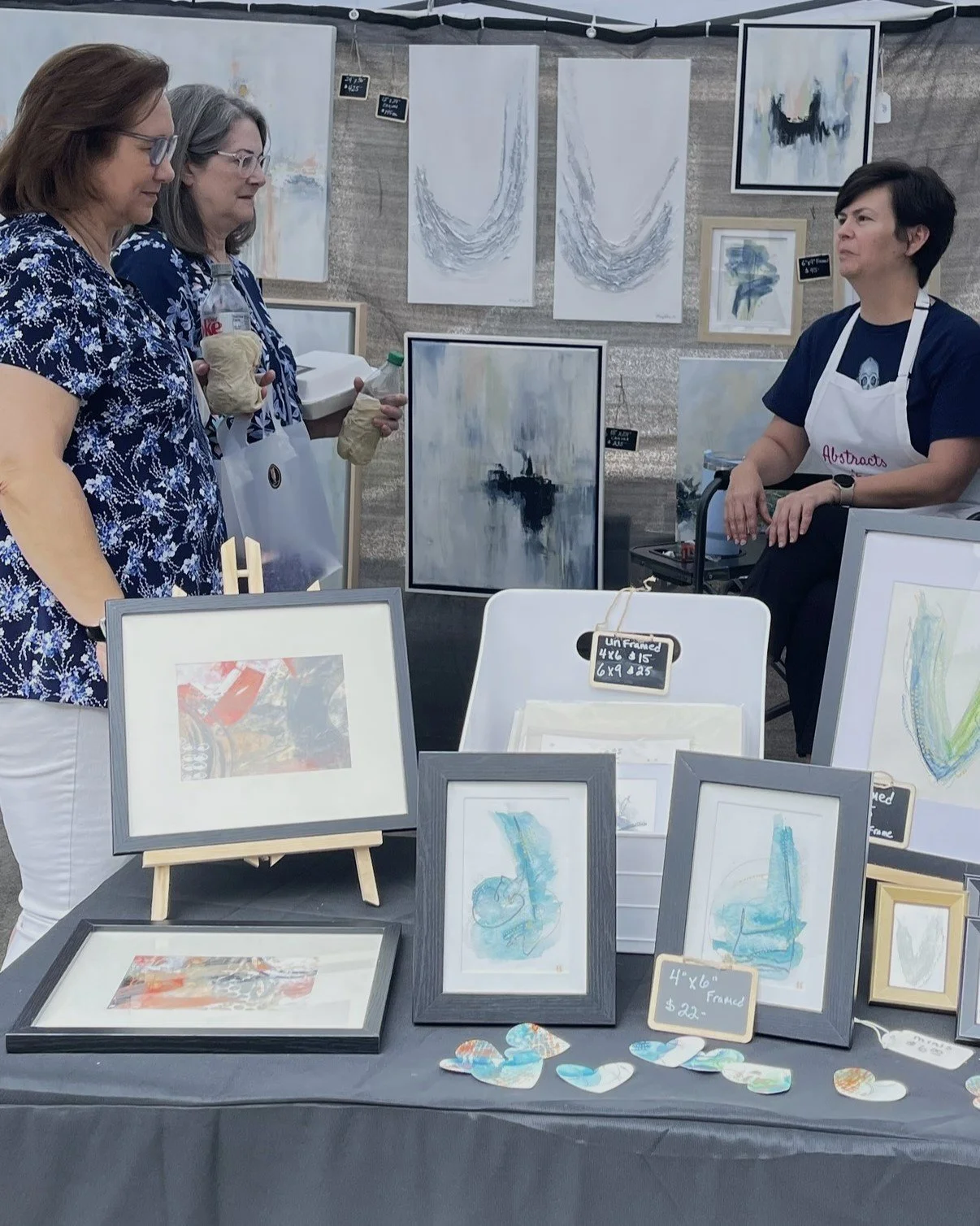 Three women viewing artwork at an art booth. The booth displays abstract watercolor paintings and framed art on a table and wall, with price tags for the framed pieces.