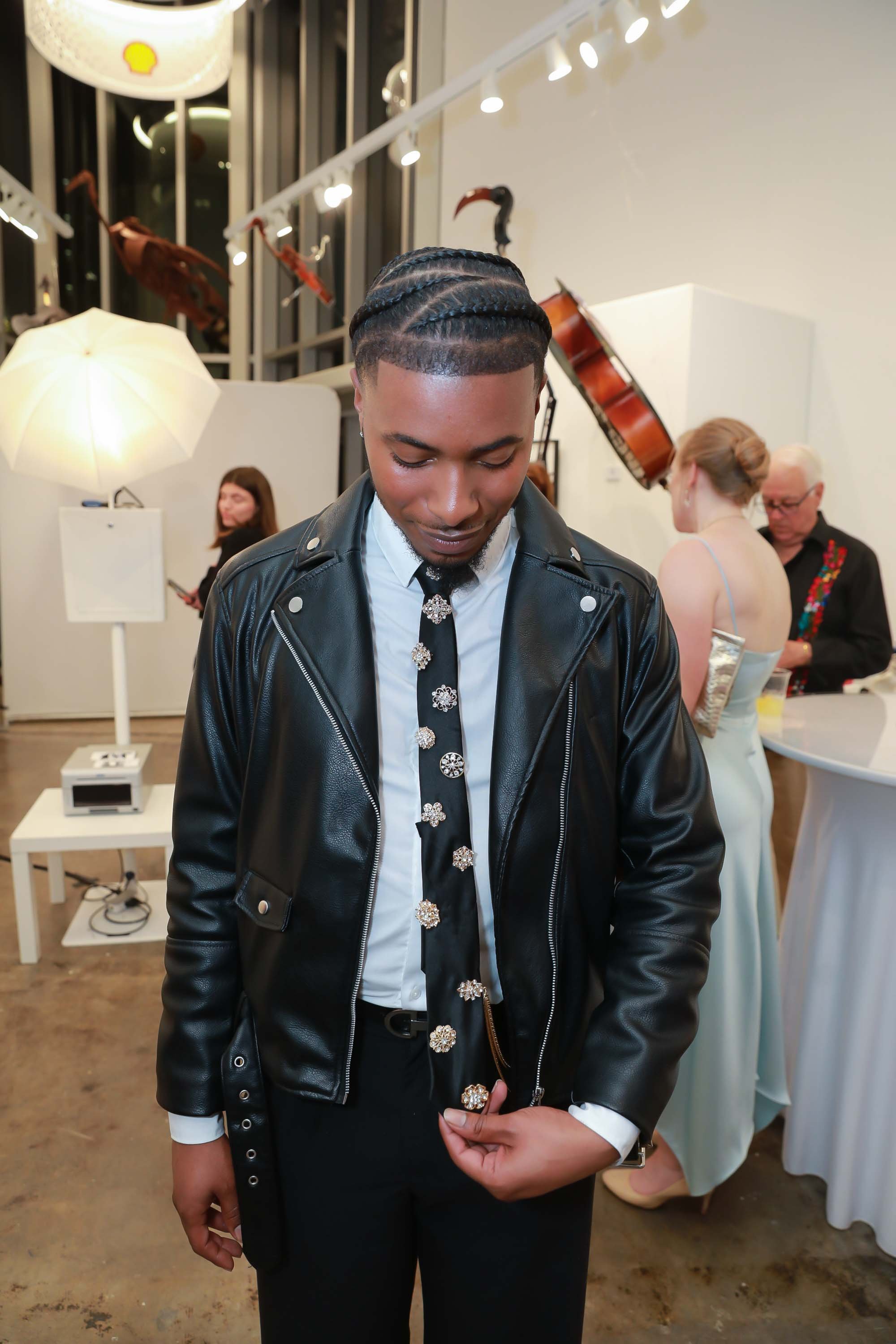 Young man with braided hairstyle wearing a leather jacket and decorative tie at an art gallery or exhibition. In the background, people are conversing and looking at art pieces, with a cello and bird sculptures on the wall, and a white umbrella-style
