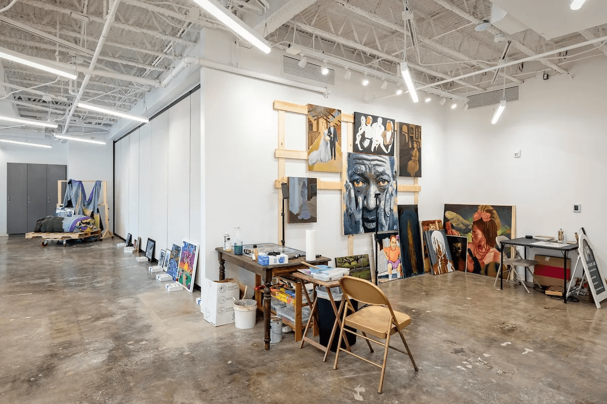 Art studio with various paintings and art supplies, including framed artworks on the floor, on tables, and a display wall, with some chairs and storage cabinets.