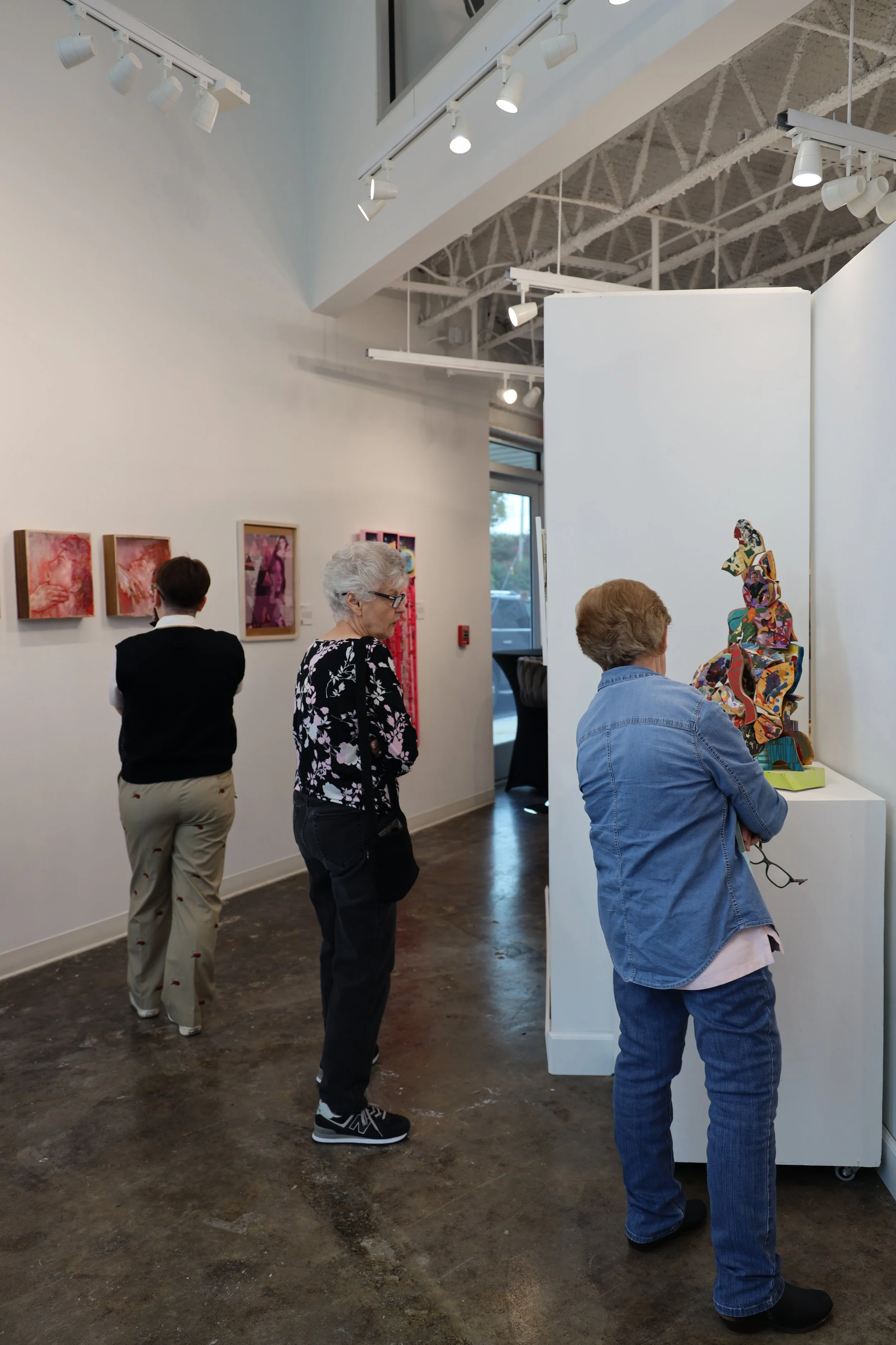 People are viewing artwork in an art gallery, with framed paintings on the wall and sculptures displayed on a white partition.