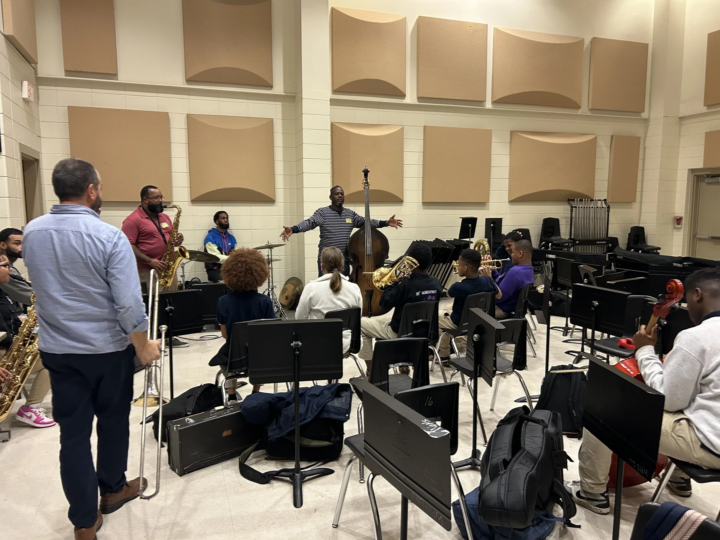 A group of musicians practicing in a music room. There are several people with musical instruments including saxophones, drums, and a double bass. One person is standing and speaking to the group with arms outstretched. The room has beige acoustic panels on the walls and black chairs.