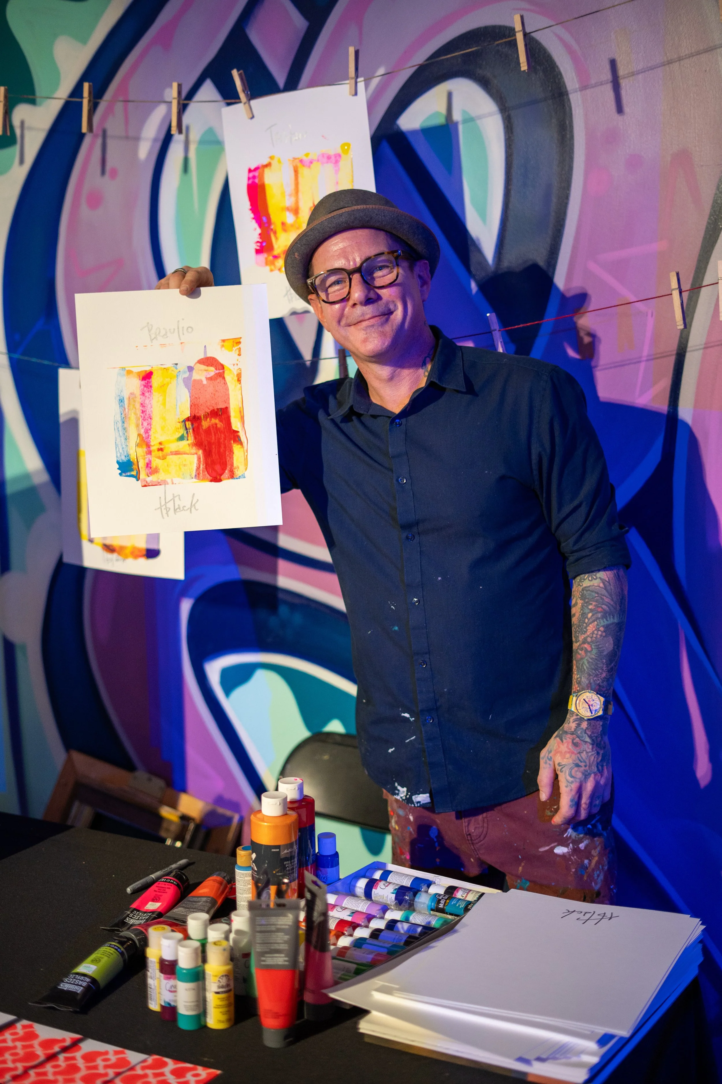A man with glasses and a hat smiling at an art studio with colorful paint bottles, paints, and papers on the table, with vibrant graffiti art on the wall in the background.