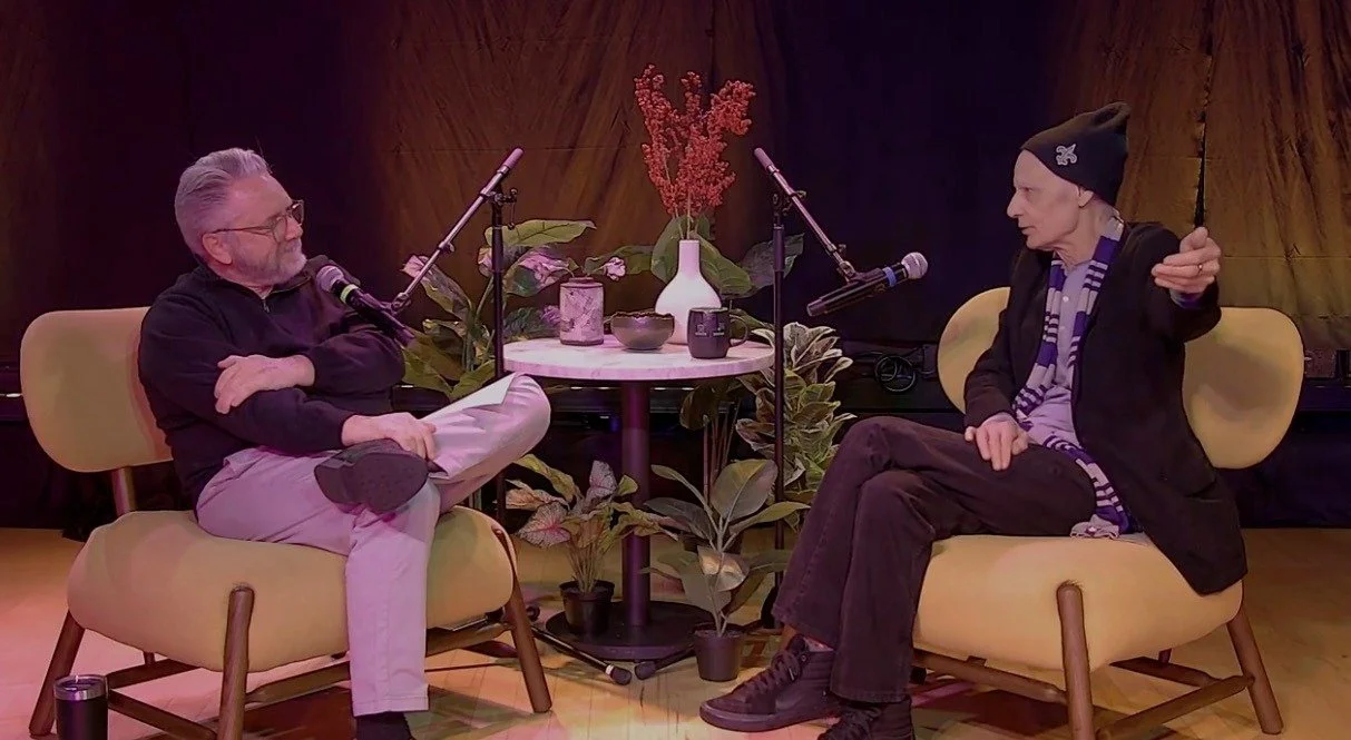 Two men having a conversation during a podcast, seated in yellow armchairs across from each other, with microphones and plants in the background.