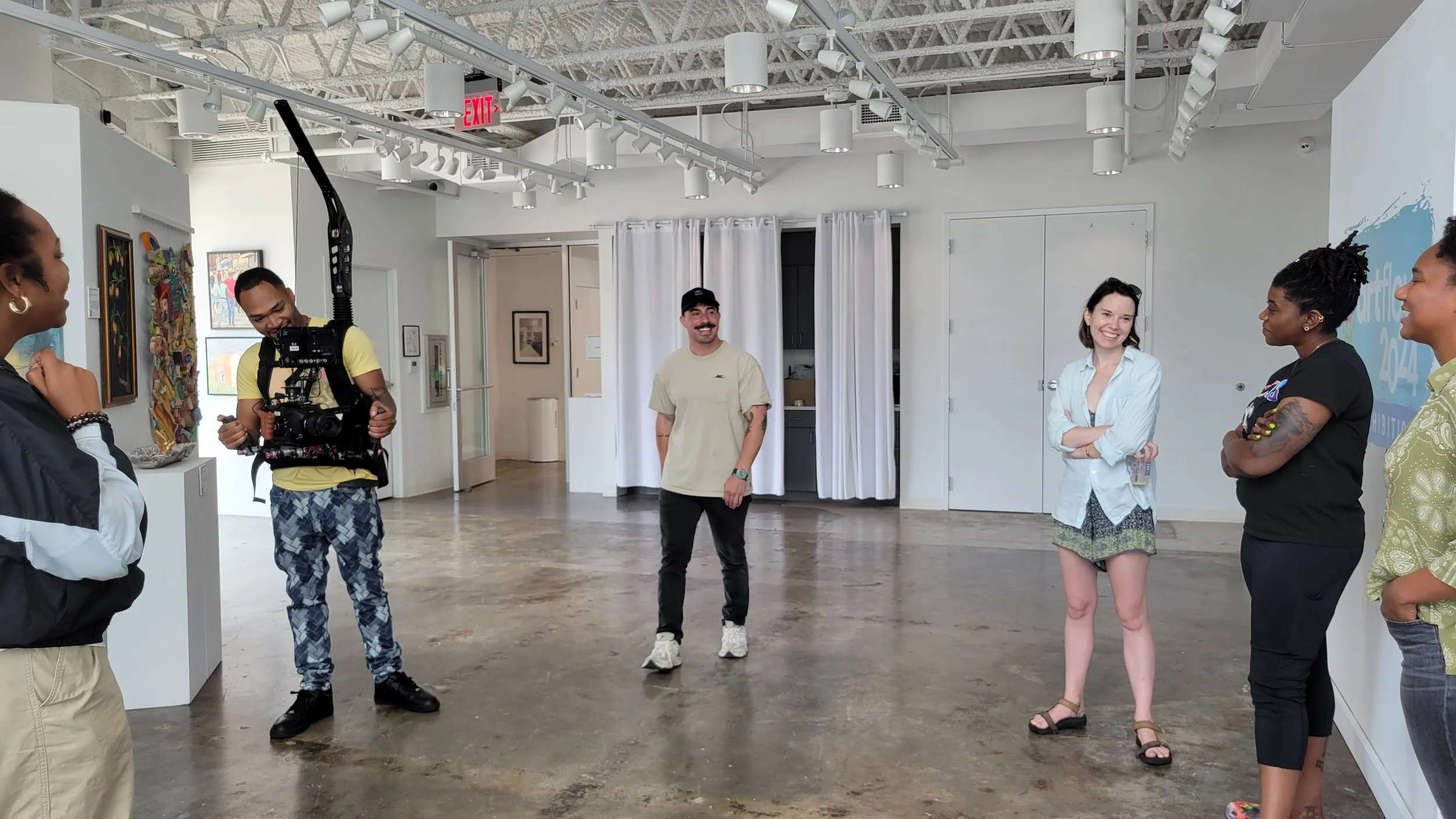 People standing in a gallery or art space, engaged in conversation, with some looking at each other and smiling. One person holds a camera, and the room has artwork on the walls, white curtains, and a ceiling with track lighting.