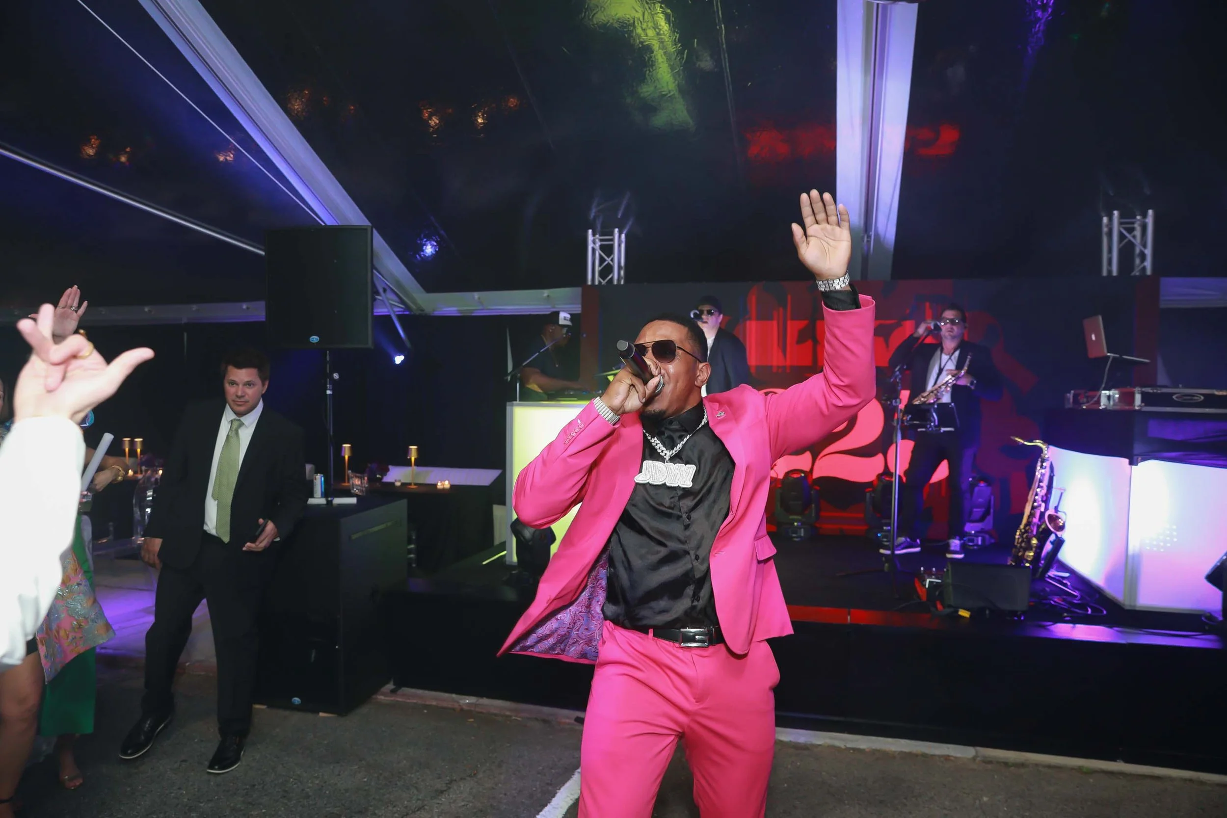 A man in a bright pink suit and black shirt performing on stage, holding a microphone, with other musicians and a DJ setup in the background at a lively indoor event.