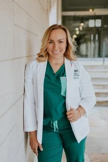 Smiling female healthcare professional in green scrubs and white lab coat standing indoors.