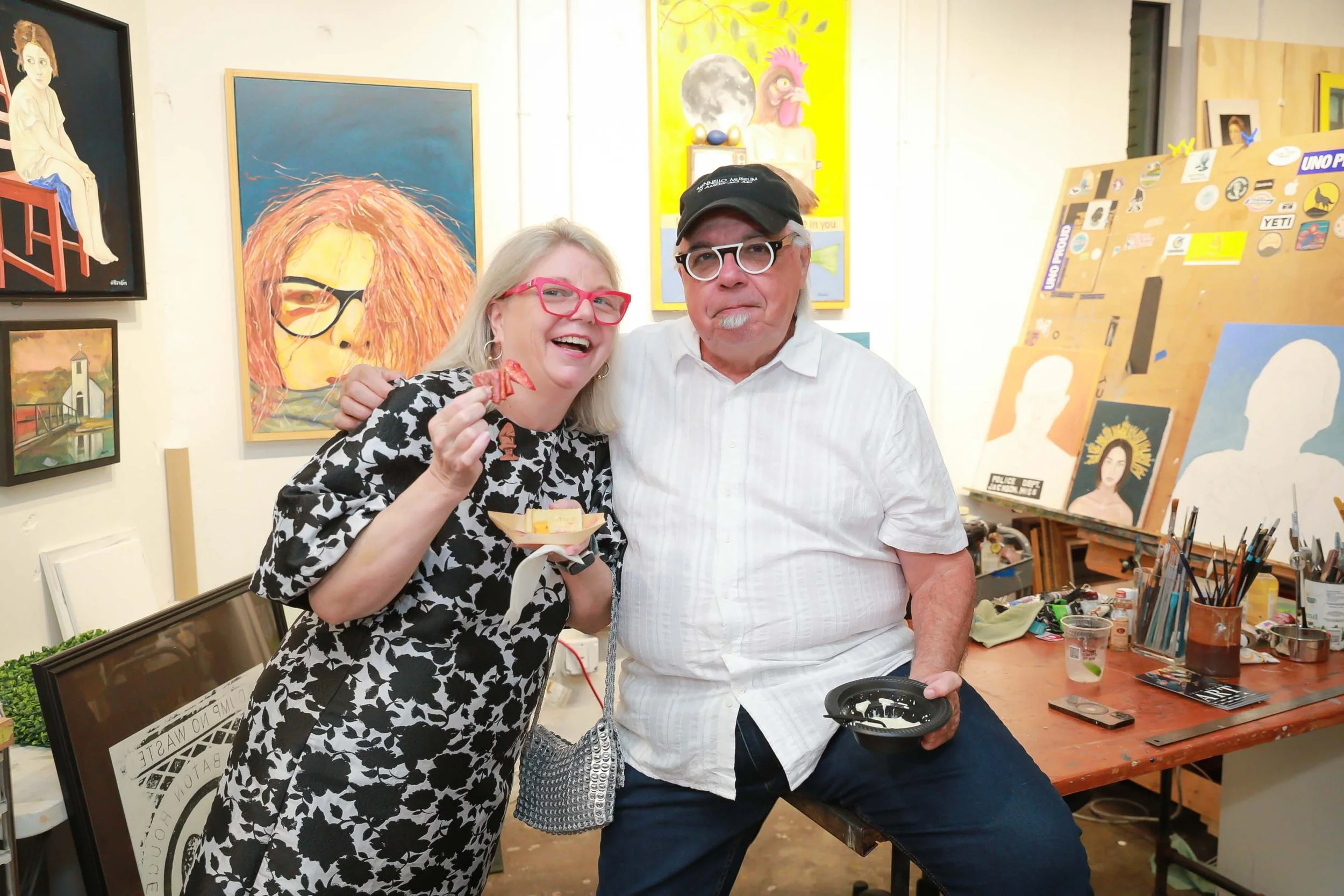 A smiling woman with blonde hair and pink glasses holding a small red object, standing next to an older man with gray hair, a beard, glasses, and a black cap. They are in an art studio with paintings on the wall and supplies on the table.