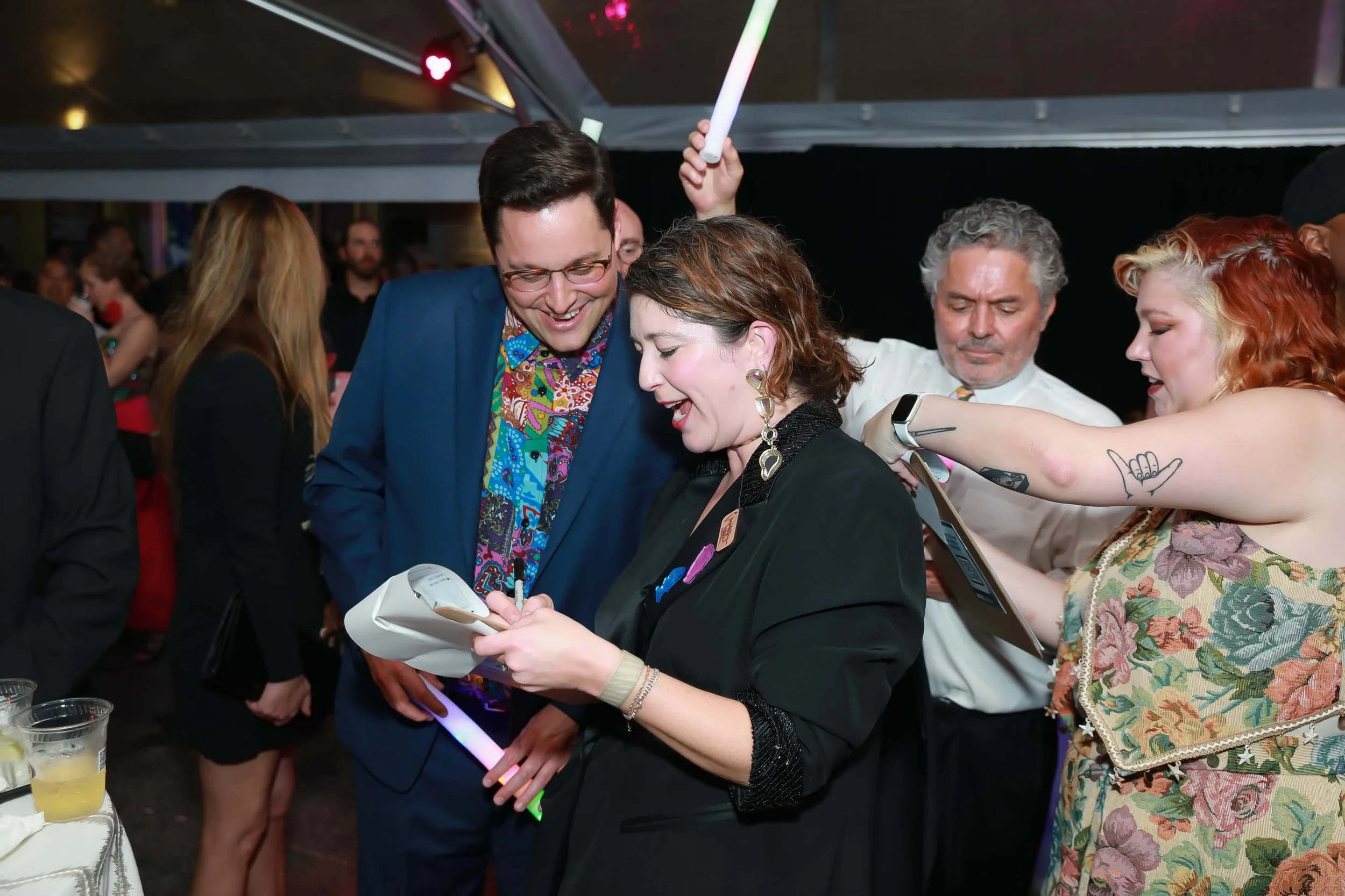 People gathered at a social event, with one woman signing an autograph for a man in a blue blazer. Others are engaged in conversation or observing the scene.