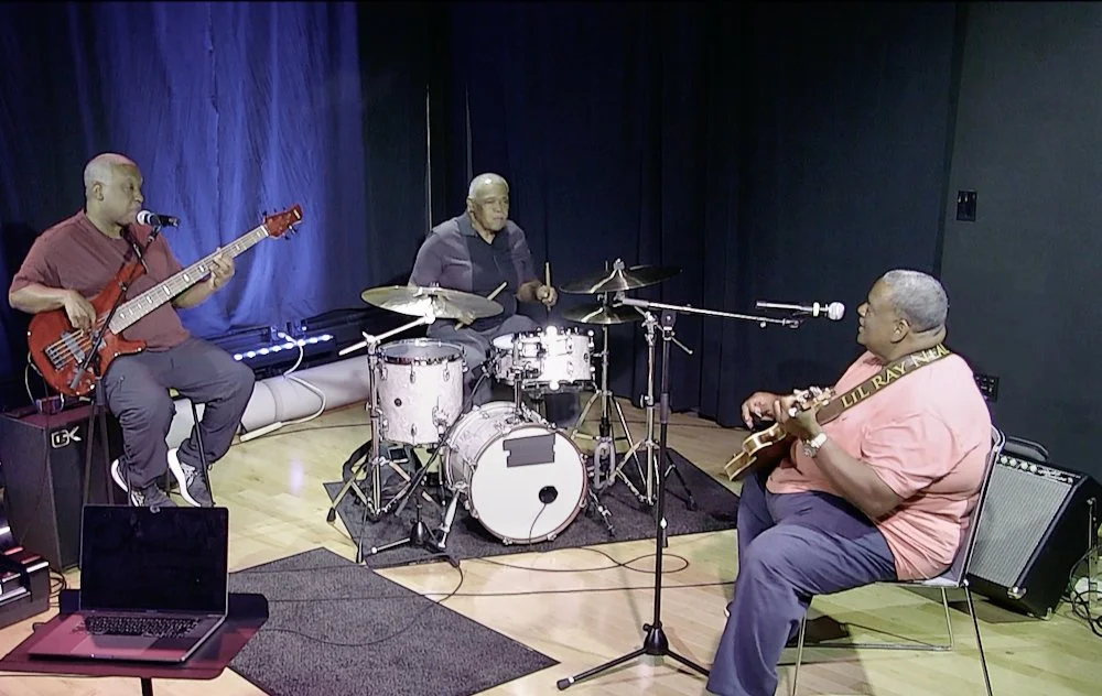Three musicians playing instruments in a recording studio: a bassist on the left, a drummer in the middle, and a guitarist on the right.