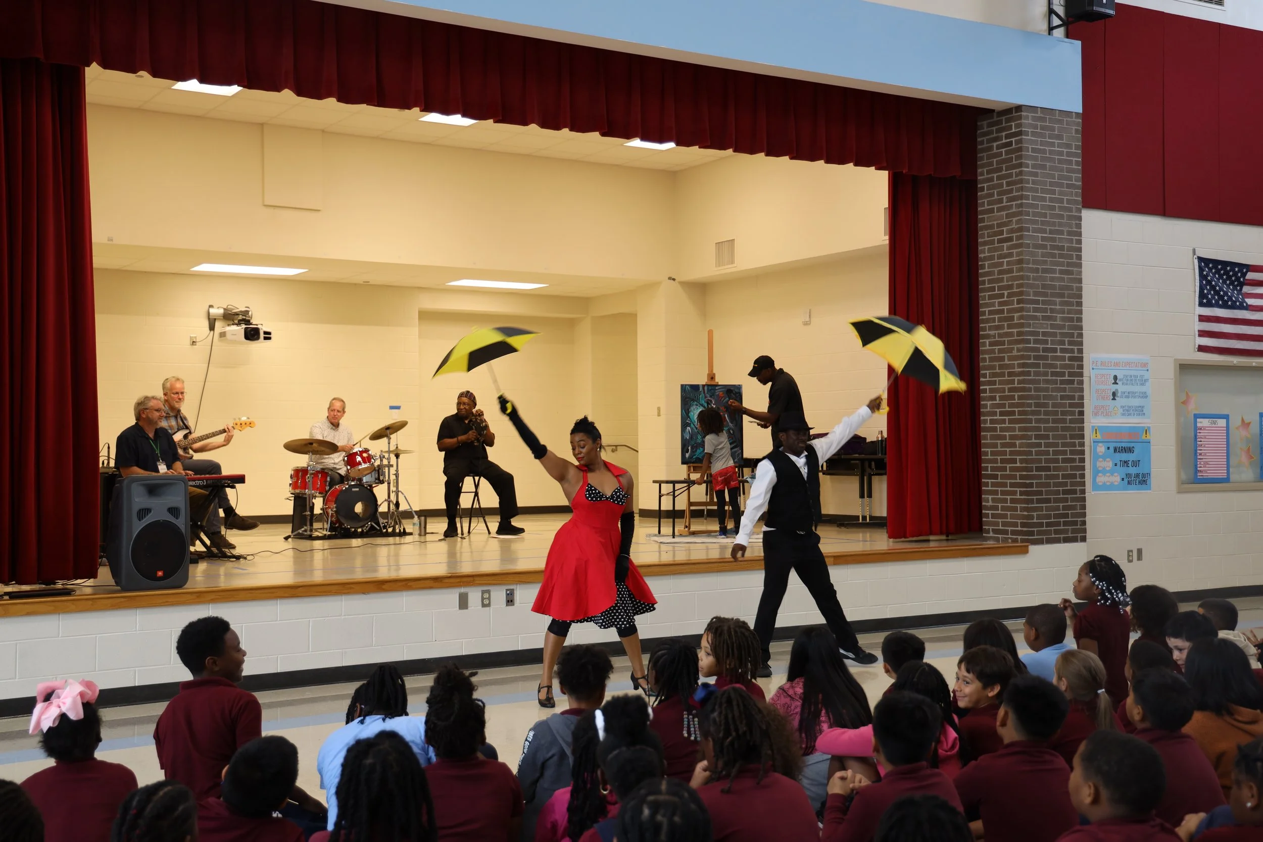 Young performers dance on stage with musicians playing instruments, while the audience, mostly children, watch from the front in a school auditorium.