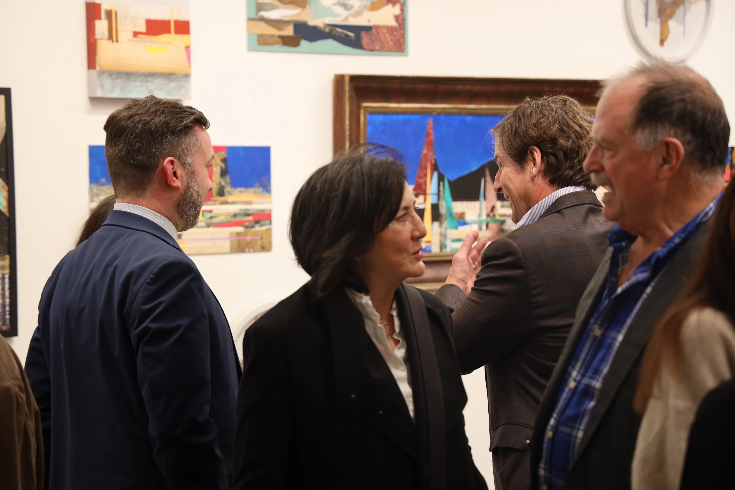 A group of five people, two men and three women, engaged in conversation at an art gallery or exhibition. The background features framed paintings and artwork on the white walls.