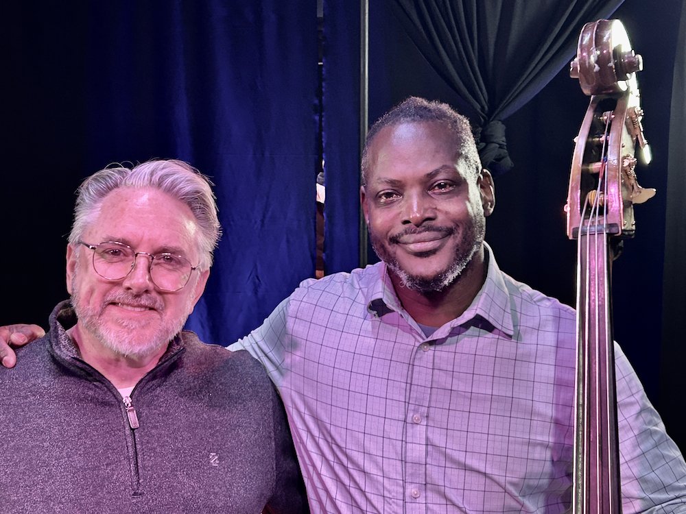 Two men smiling, one with gray hair and glasses, wearing a gray sweater, and the other with a beard and short hair, holding a double bass, against a dark blue curtain background.
