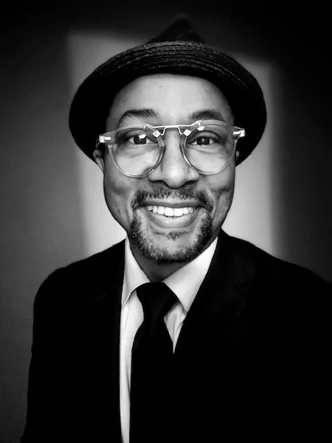 A black-and-white photo of a smiling man wearing a hat, large glasses, a suit, and a tie.