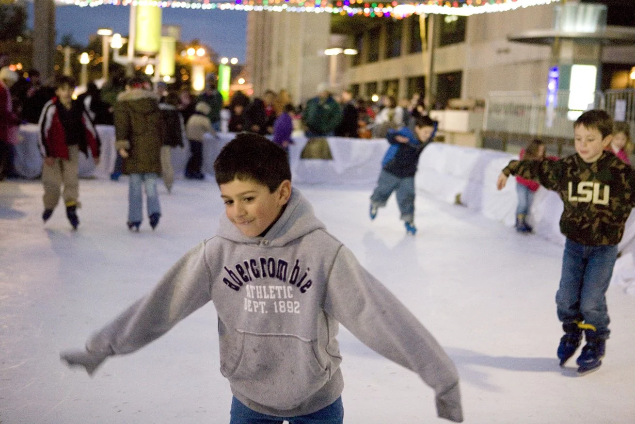 Children ice skating outdoors at night, with string lights overhead and a busy urban background.