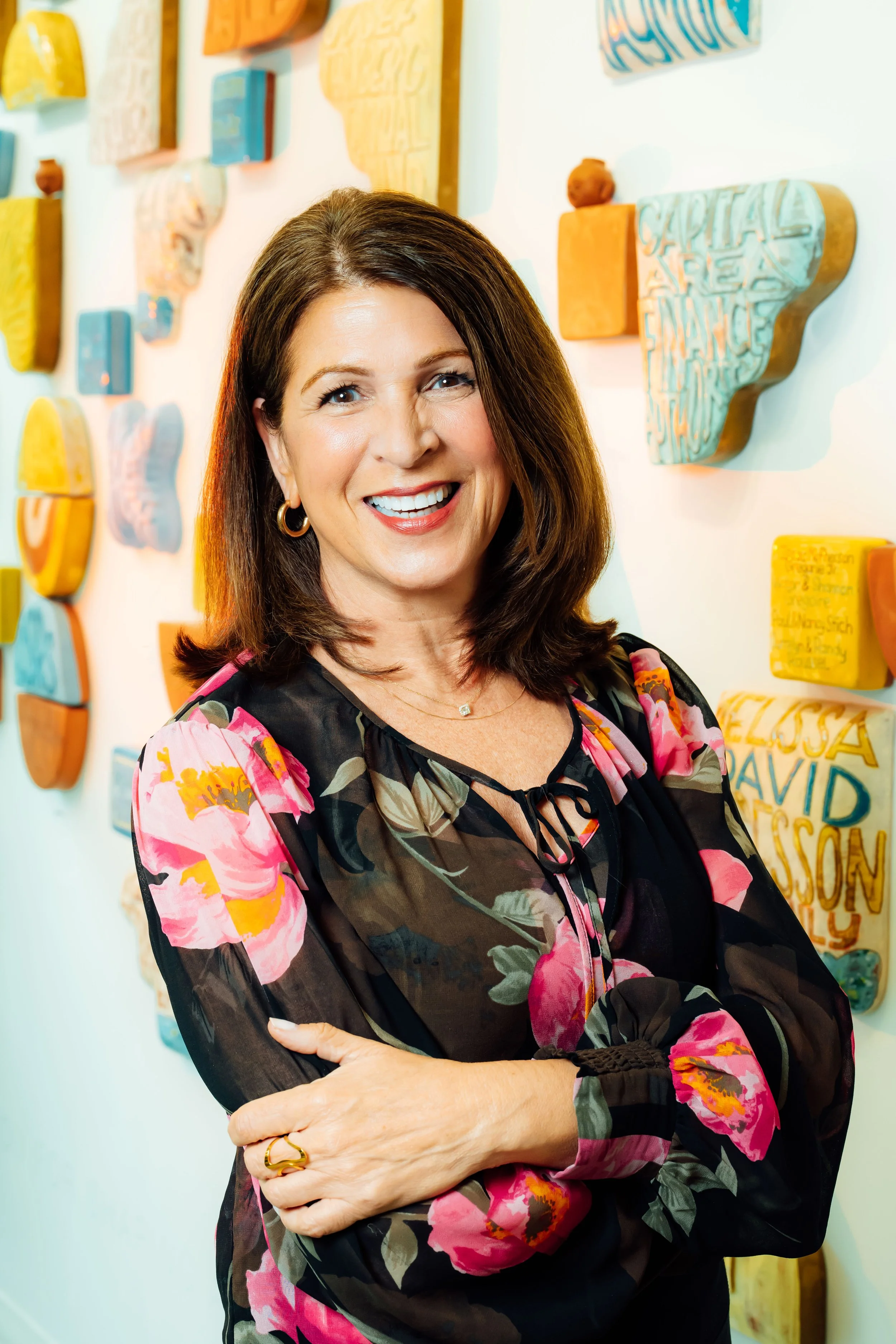 A woman with shoulder-length brown hair wearing a black blouse with pink floral sleeves, gold hoop earrings, and a gold ring, standing with arms crossed in front of a wall decorated with colorful, textured art pieces.