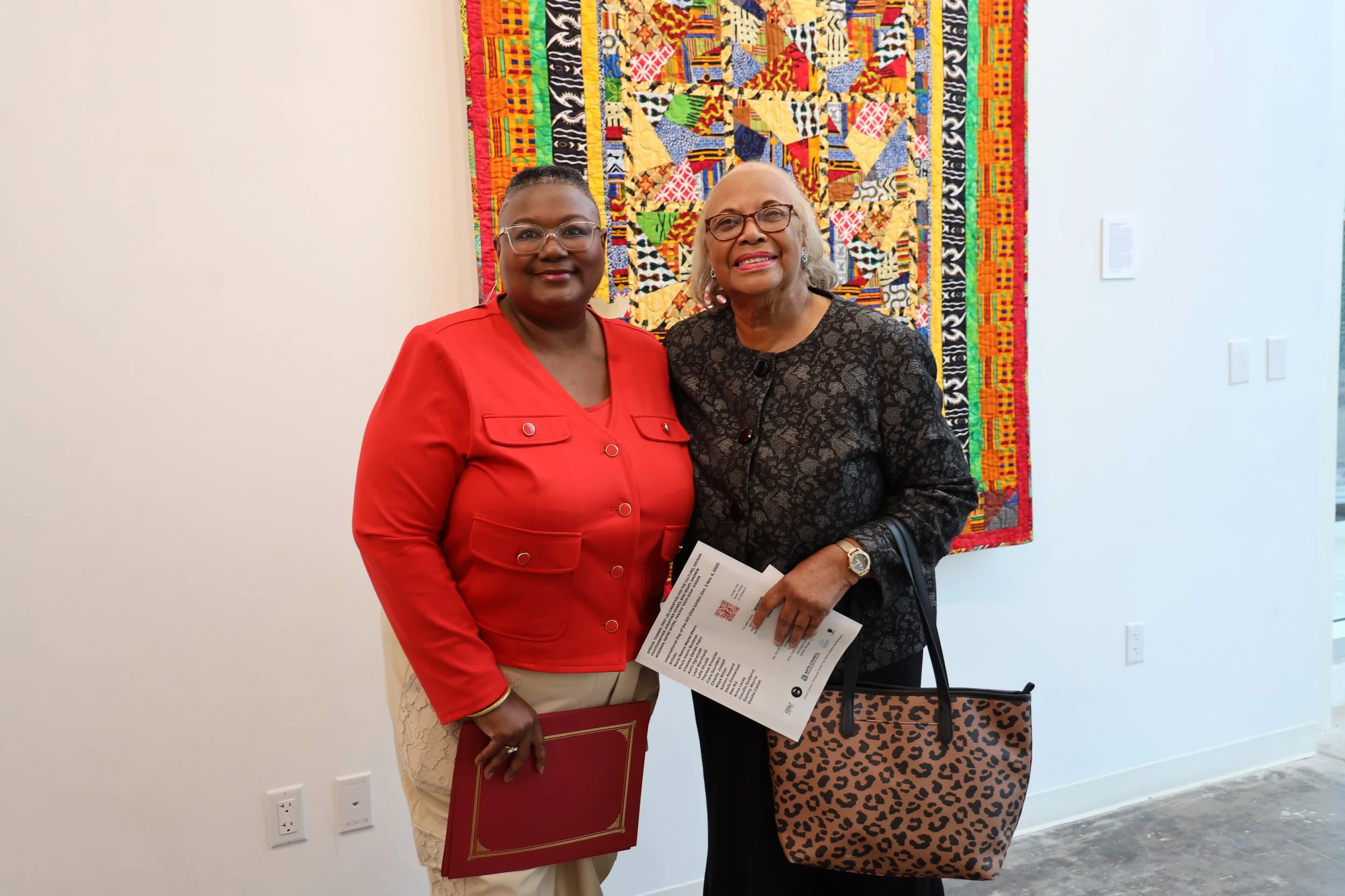 Two women standing in front of a colorful quilt; one woman is wearing glasses and a red dress, holding a folder, and the other woman, with gray hair, glasses, and wearing a black lace top with animal print skirt, is holding a pamphlet.