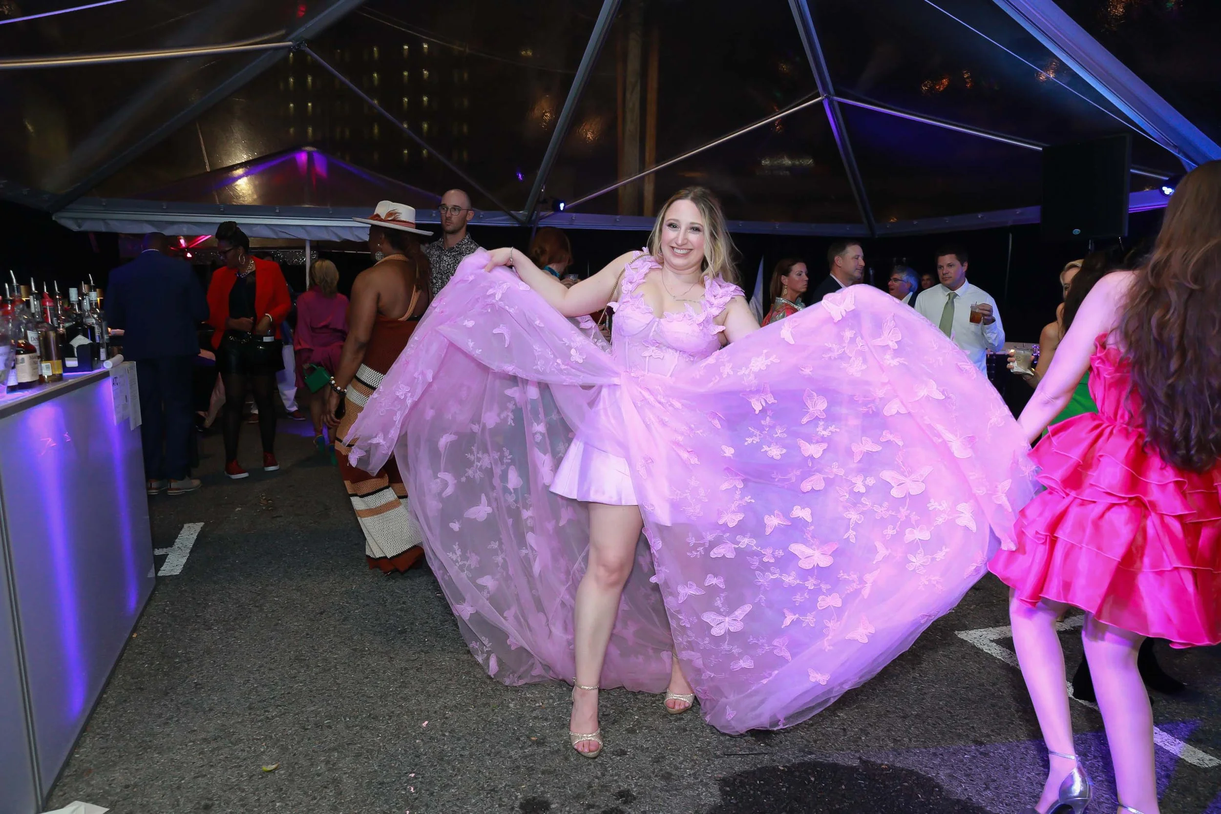 A woman in a pink, butterfly-patterned ball gown is smiling and posing on a dance floor at an event or party, with people mingling in the background under a transparent tent with purple lighting.