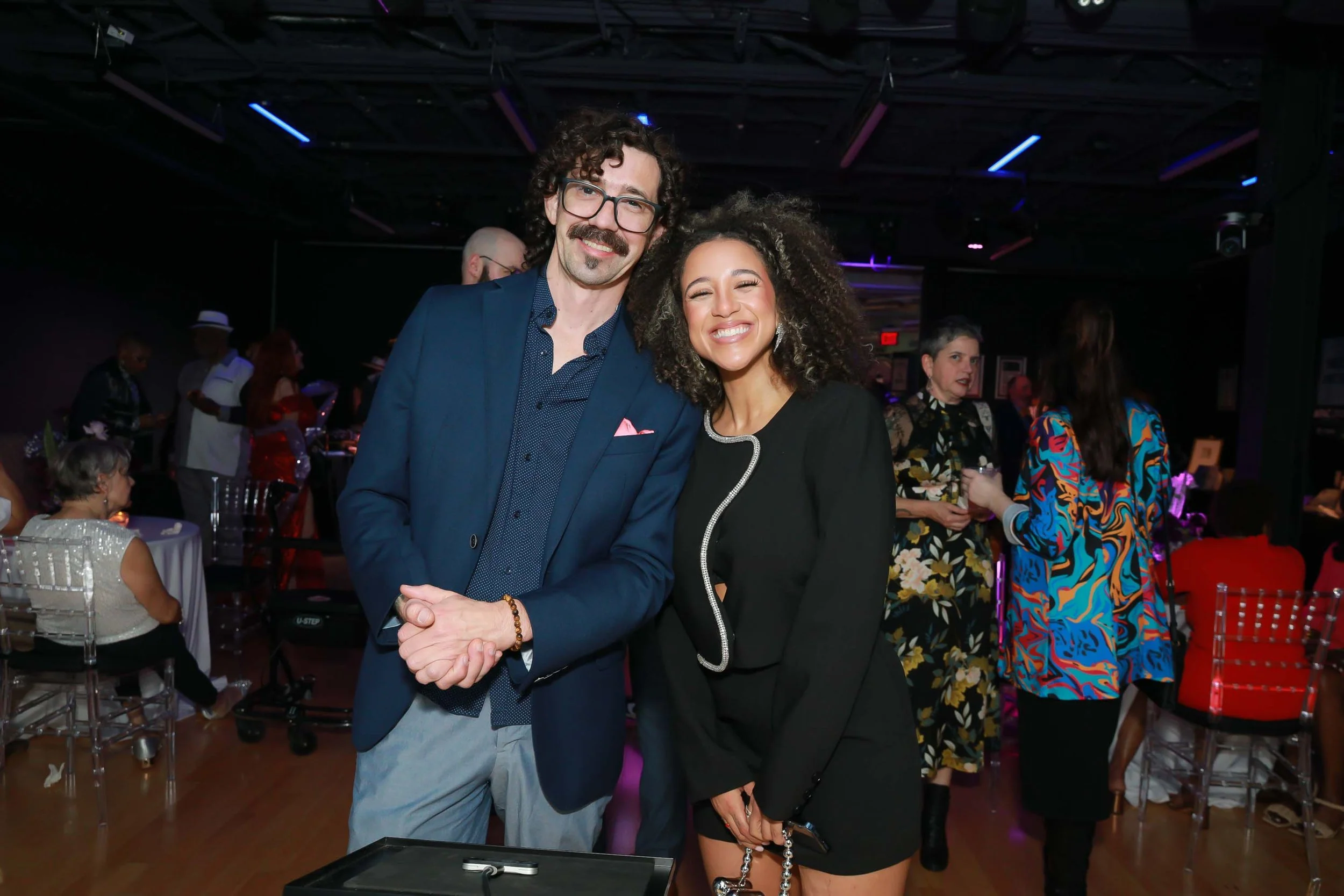 A man with curly hair, glasses, and a mustache dressed in a blue blazer and light gray pants shaking hands with a woman with curly hair in a black blazer at a social event. In the background, other people are mingling, some seated at tables, and some