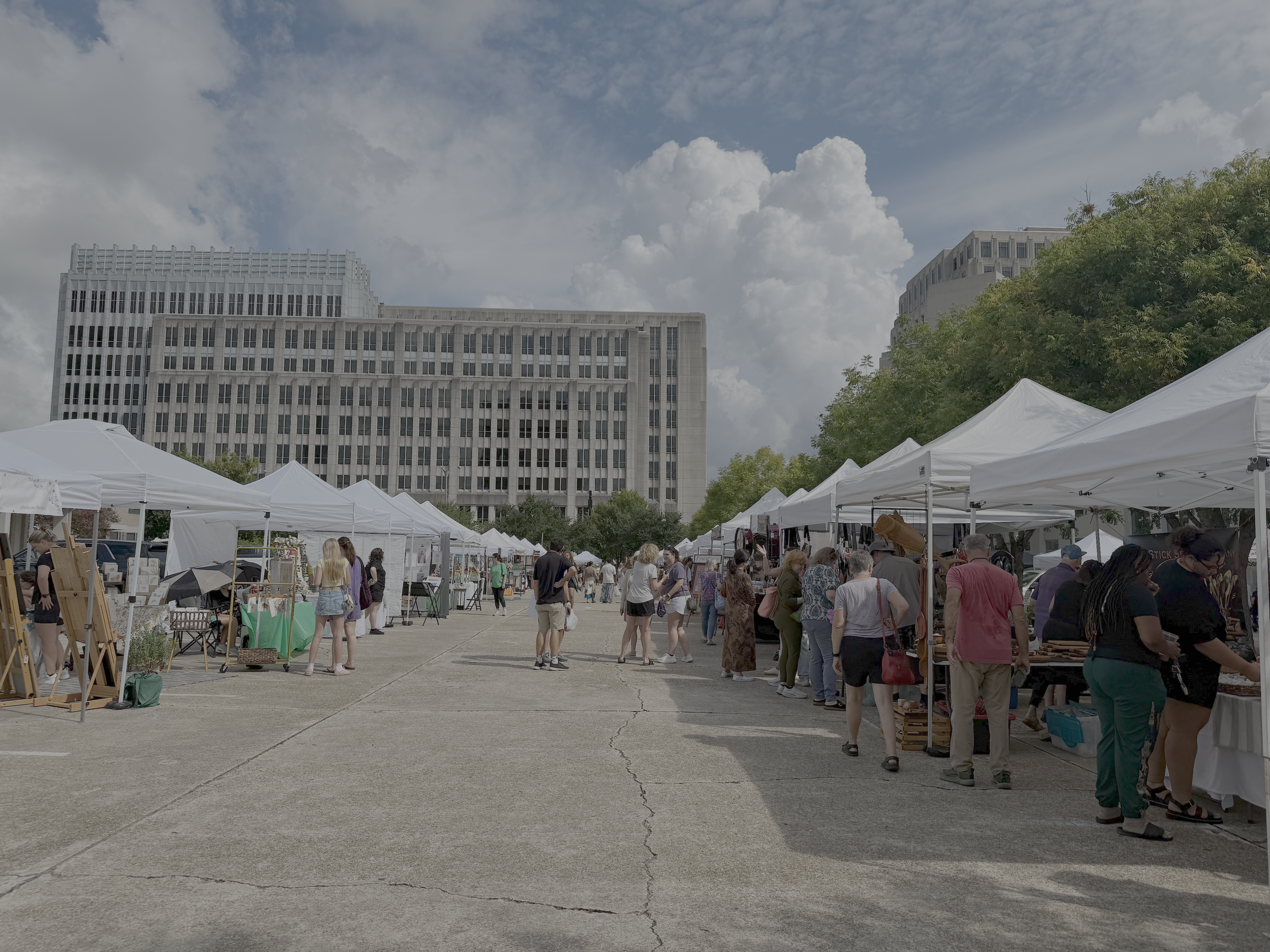 Outdoor street fair with vendor booths, people browsing, under white tents, with modern high-rise buildings and trees in the background on a sunny day.