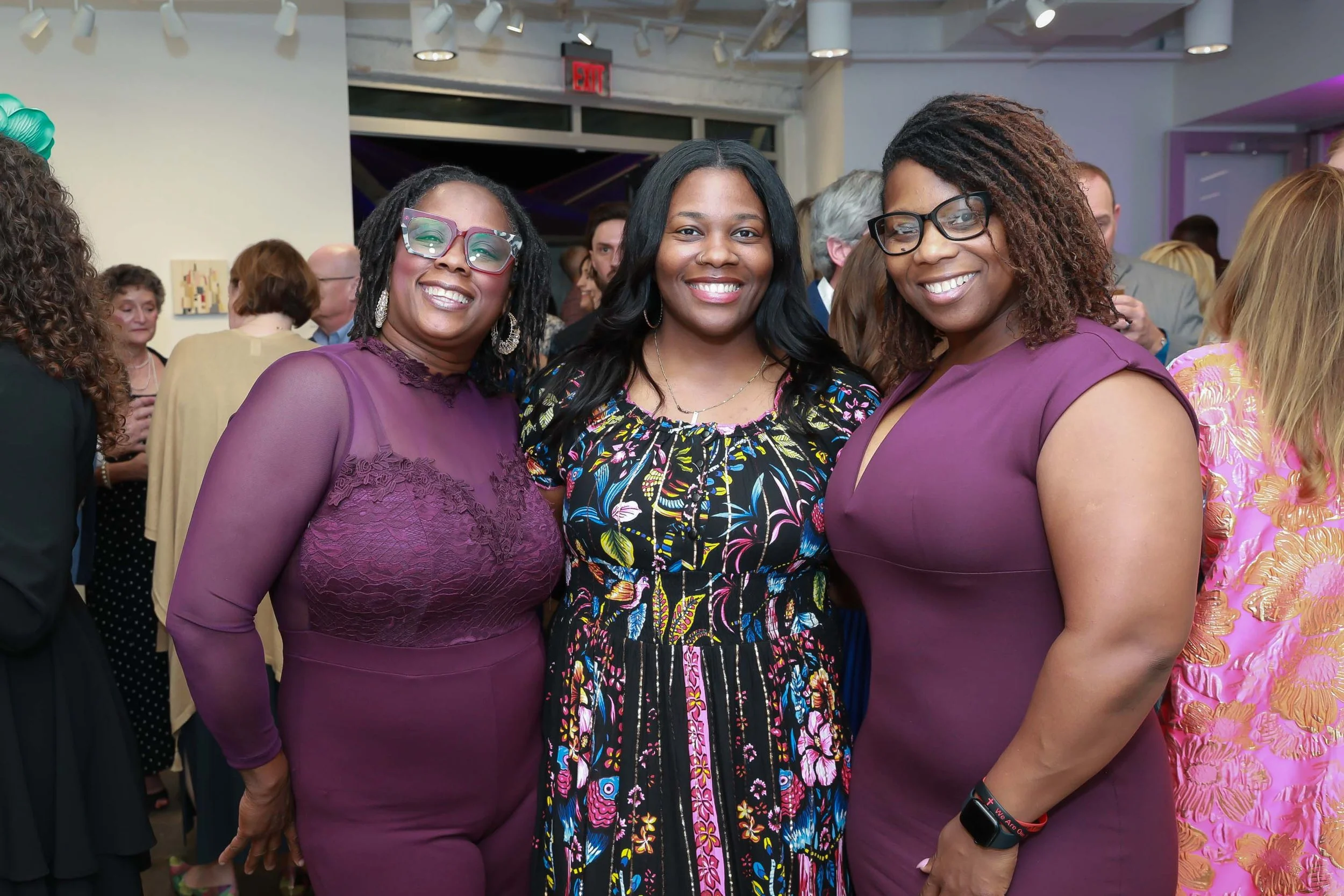 Three women smiling at an indoor event, standing close together, with a crowd of people in the background.