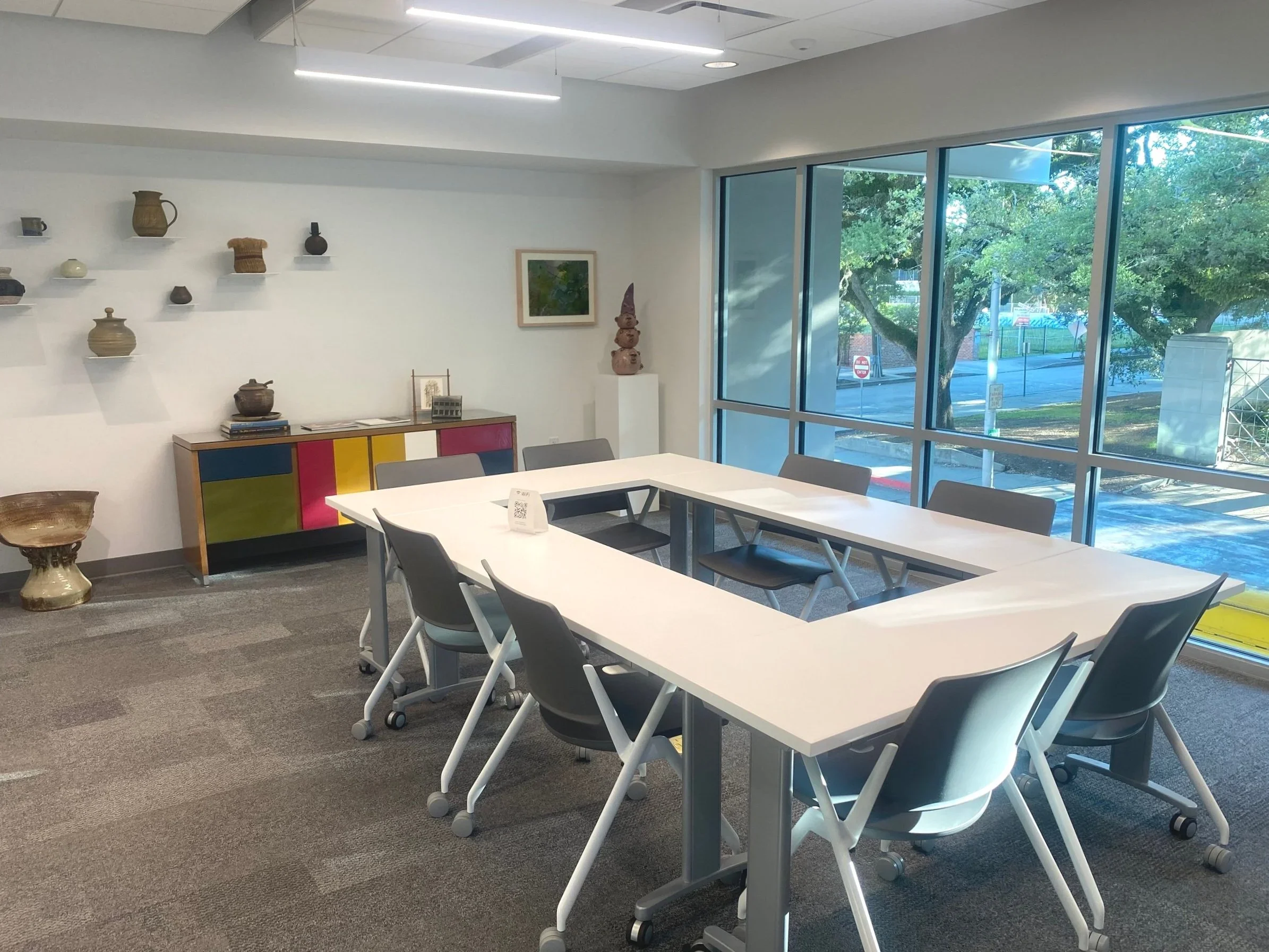 An indoor meeting room with white tables and gray chairs, wall having decorative pottery and a framed picture, large windows showing trees outside, and colorful cabinet against the wall.