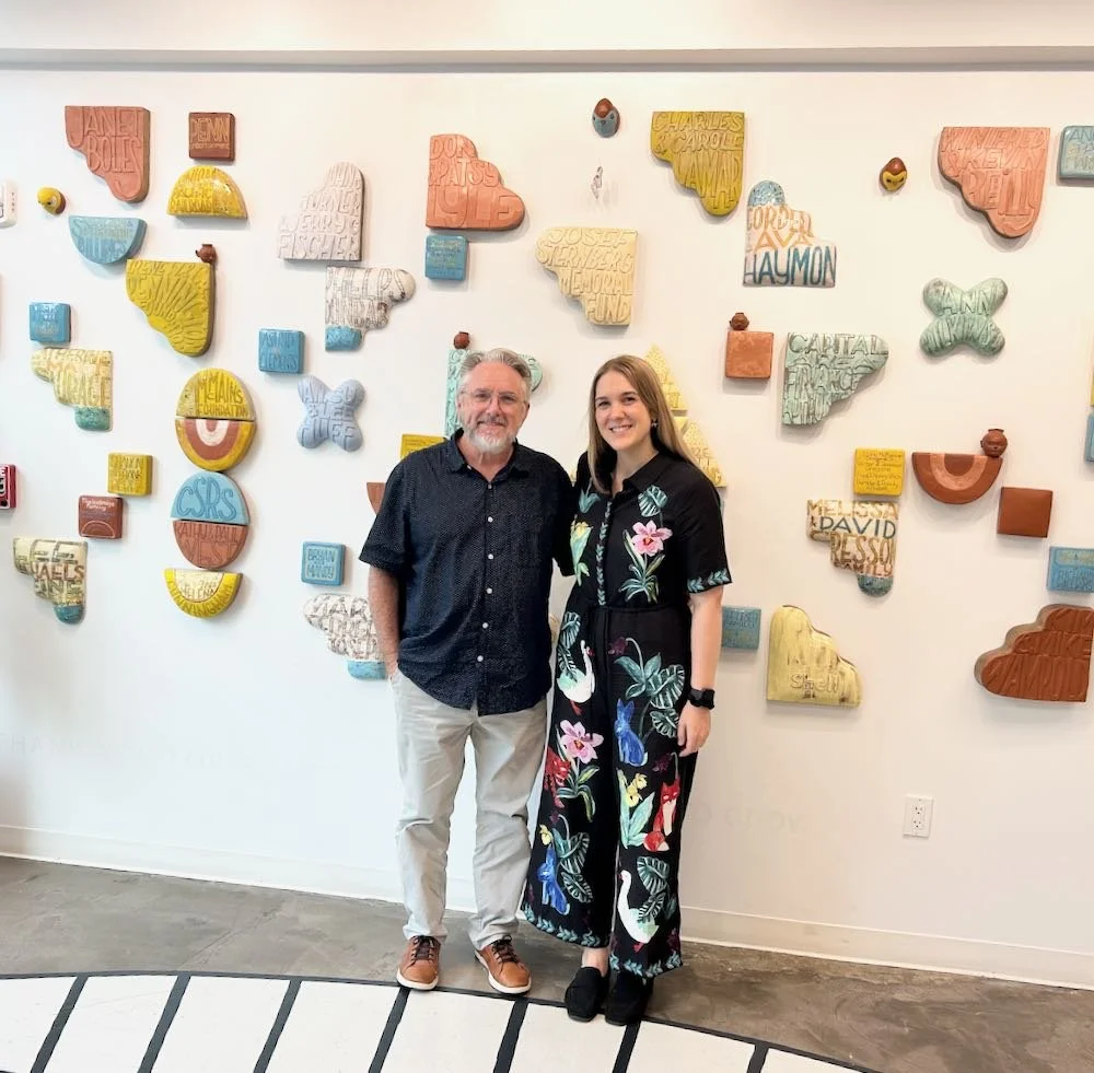 A man and a woman standing side by side in front of a colorful wall art installation. The wall features various wall-mounted blocks and shapes with text and patterns in pastel colors.