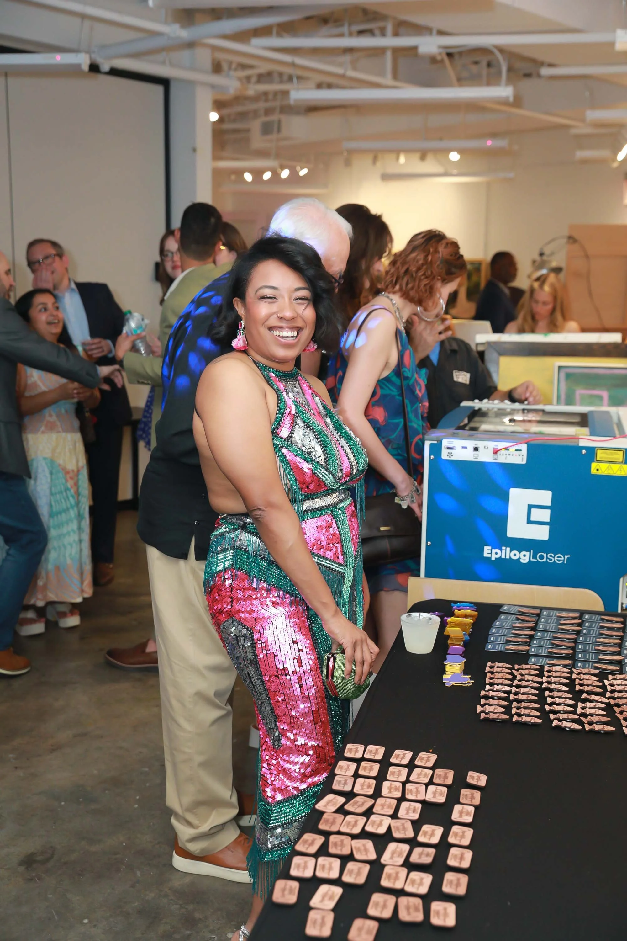 A woman in a colorful, sequined dress with pink and green, smiling at the camera, standing at a table with small tiles in an indoor event with many people in background.