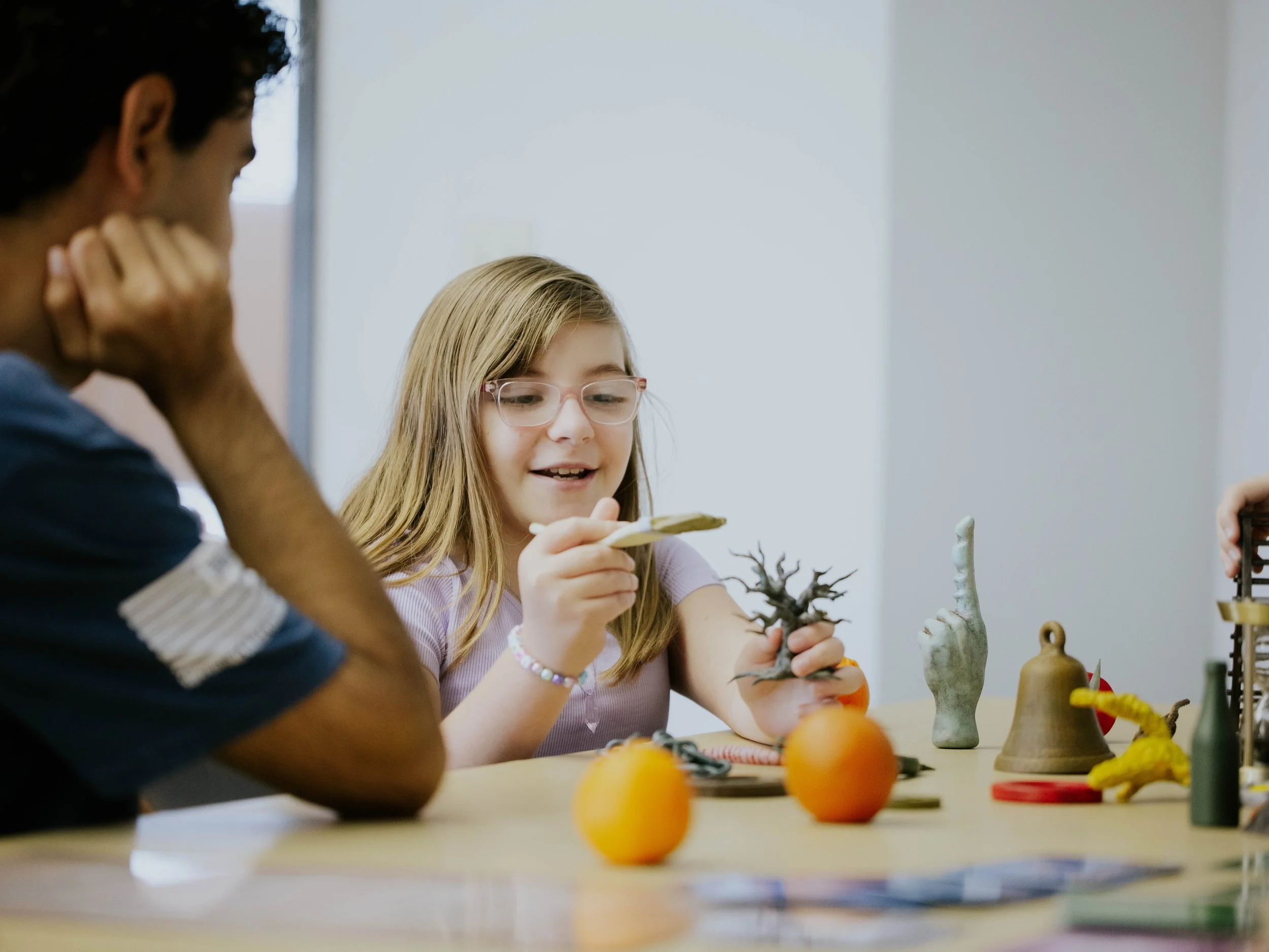 A young girl with glasses and long blonde hair sitting at a table, holding a small piece of paper and smiling. Two oranges and various decorative items are on the table. An adult with dark curly hair and dark skin is sitting next to her, resting their face on their hand, partially visible. The background is plain and white.