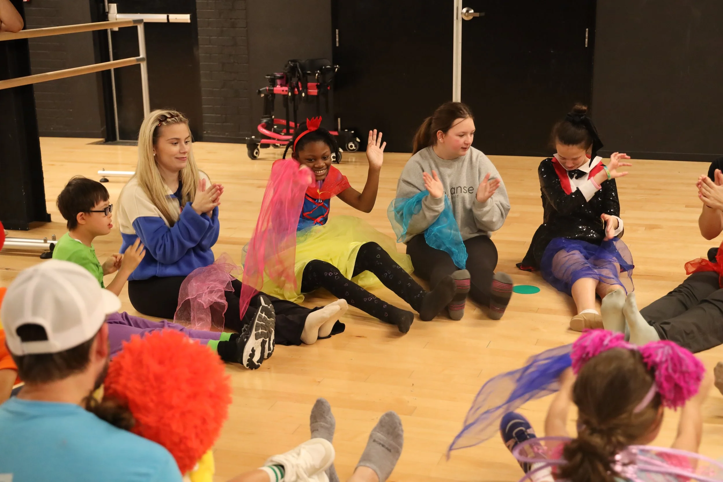 Children and a young woman sitting on the floor in a dance studio, engaging in a playful activity, with some children wearing colorful tutus and costumes.