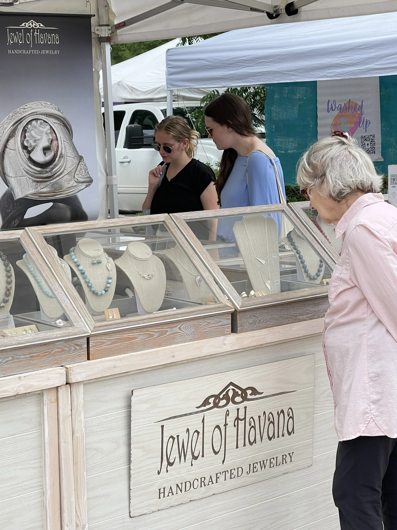 A jewelry booth at an outdoor market with a sign that reads "Jewel of Havana, Handcrafted Jewelry." Several necklaces are displayed in glass cases, and three women are browsing the jewelry, with one older woman in a pink shirt and two younger women w