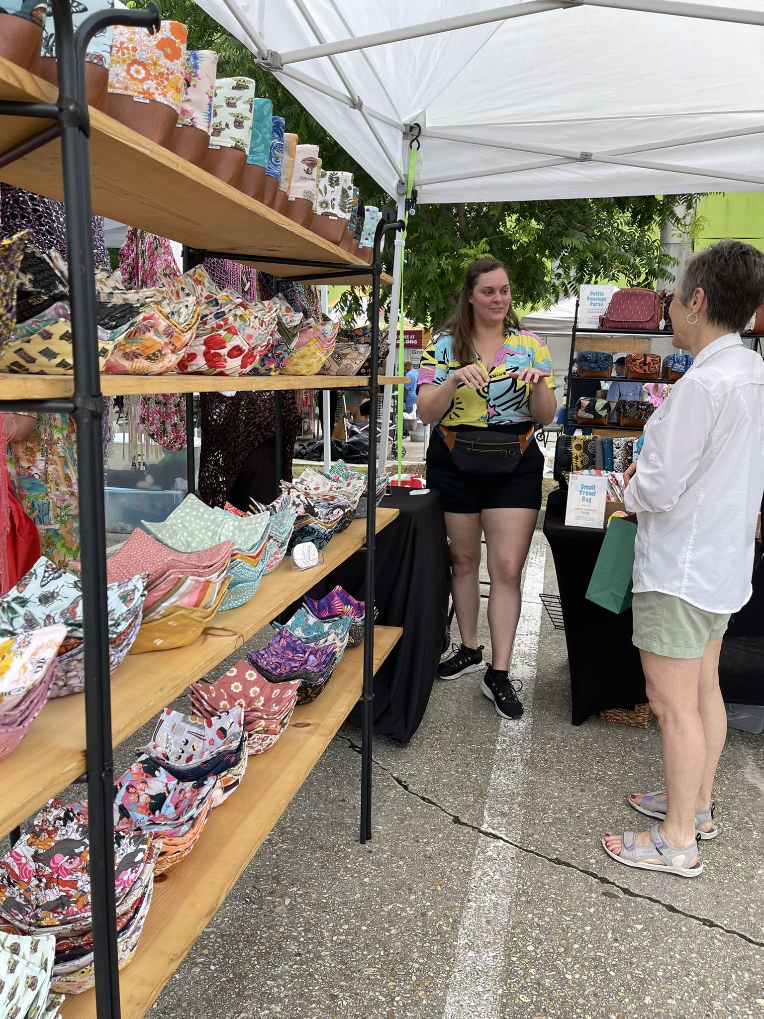 A woman and an older woman are conversing at an outdoor market stall. The stall displays various colorful fabric bags and pouches on shelves under a white canopy. The women are standing on a paved surface surrounded by other market stalls and greener