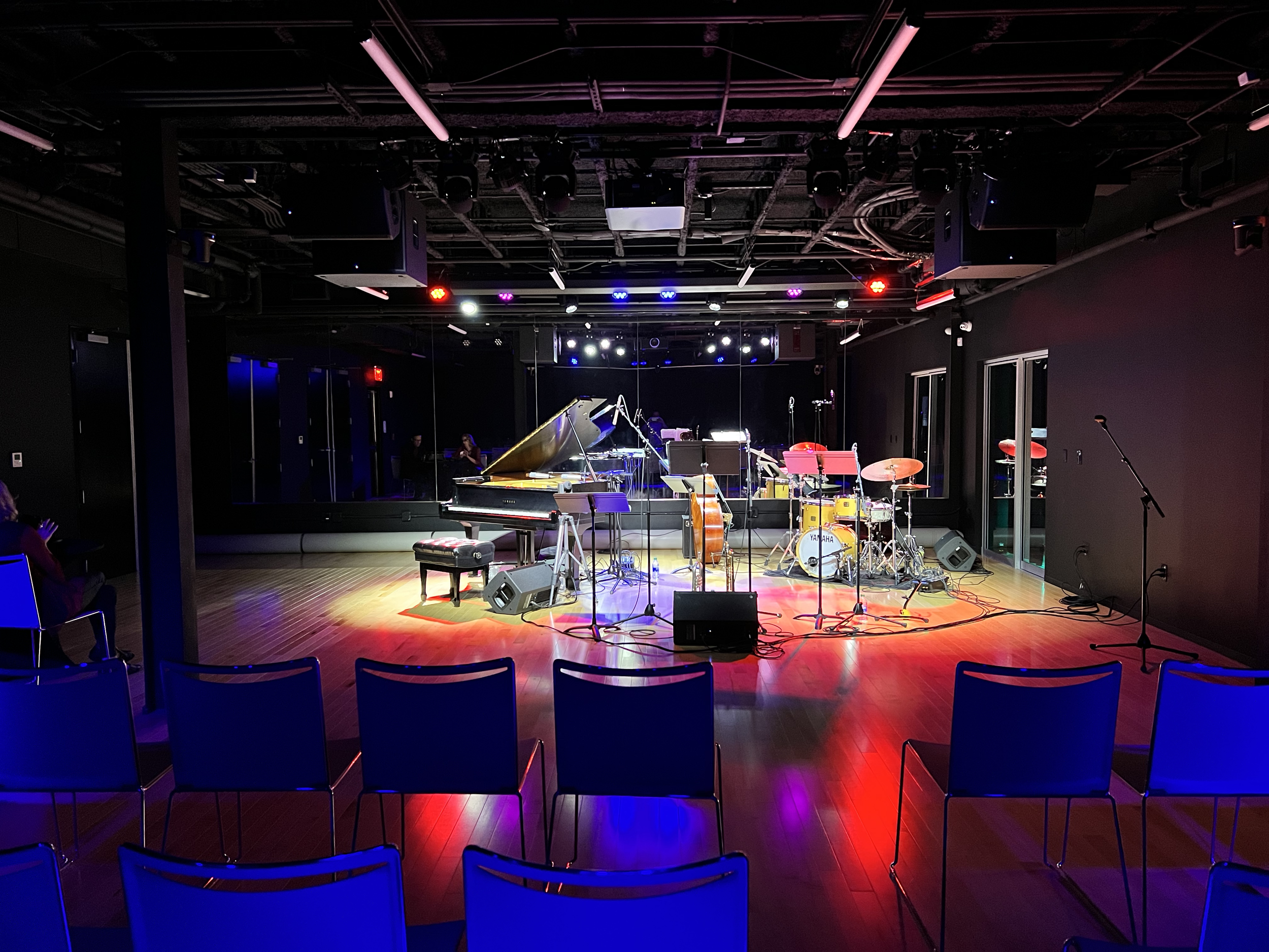A music stage with instruments including a grand piano, drums, and microphones, illuminated by colorful stage lighting, and seated audience chairs in the foreground.