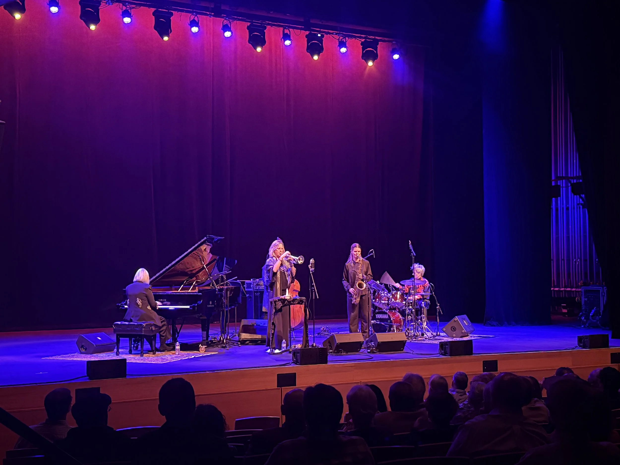 A jazz band performing on stage in a theater with purple and blue stage lighting. Musicians include a pianist, trumpet player, saxophonist, and drummer, with an audience seated and watching.