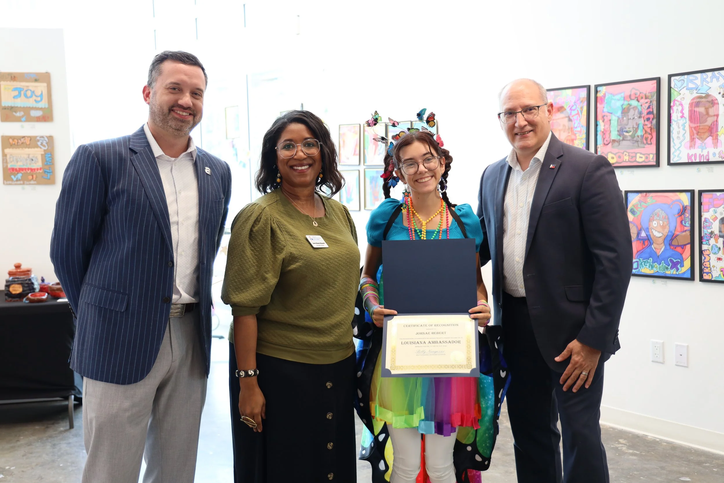 Group of five people at an art gallery, with a young girl holding a certificate, surrounded by colorful artwork.
