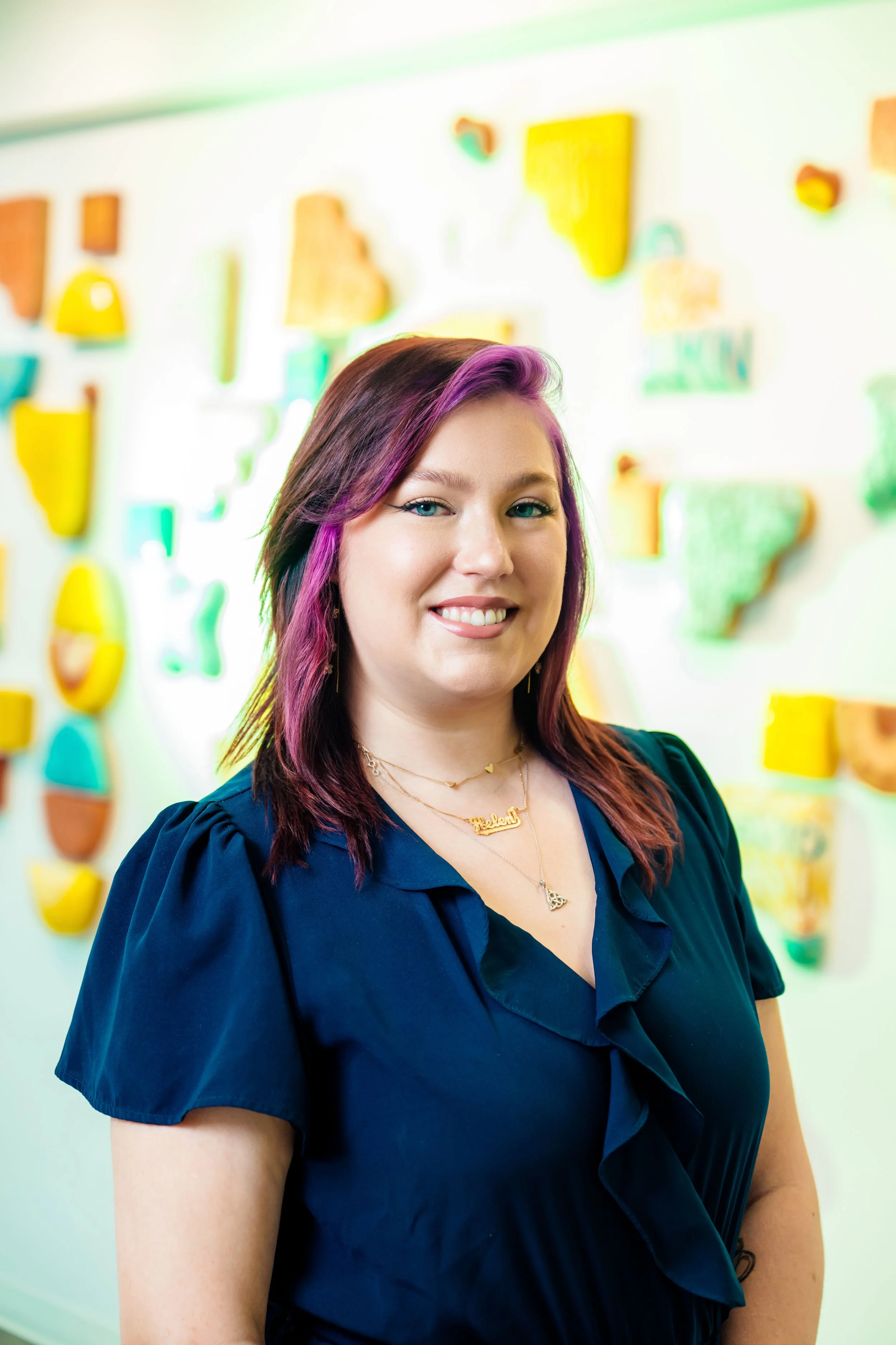 A woman with purple and brown hair smiling in front of a colorful wall display.