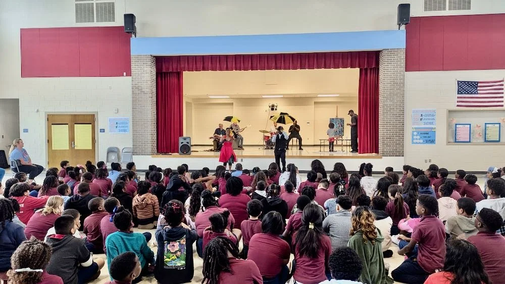 School assembly with students watching a stage performance, featuring musicians with umbrellas and a woman in a pink dress.