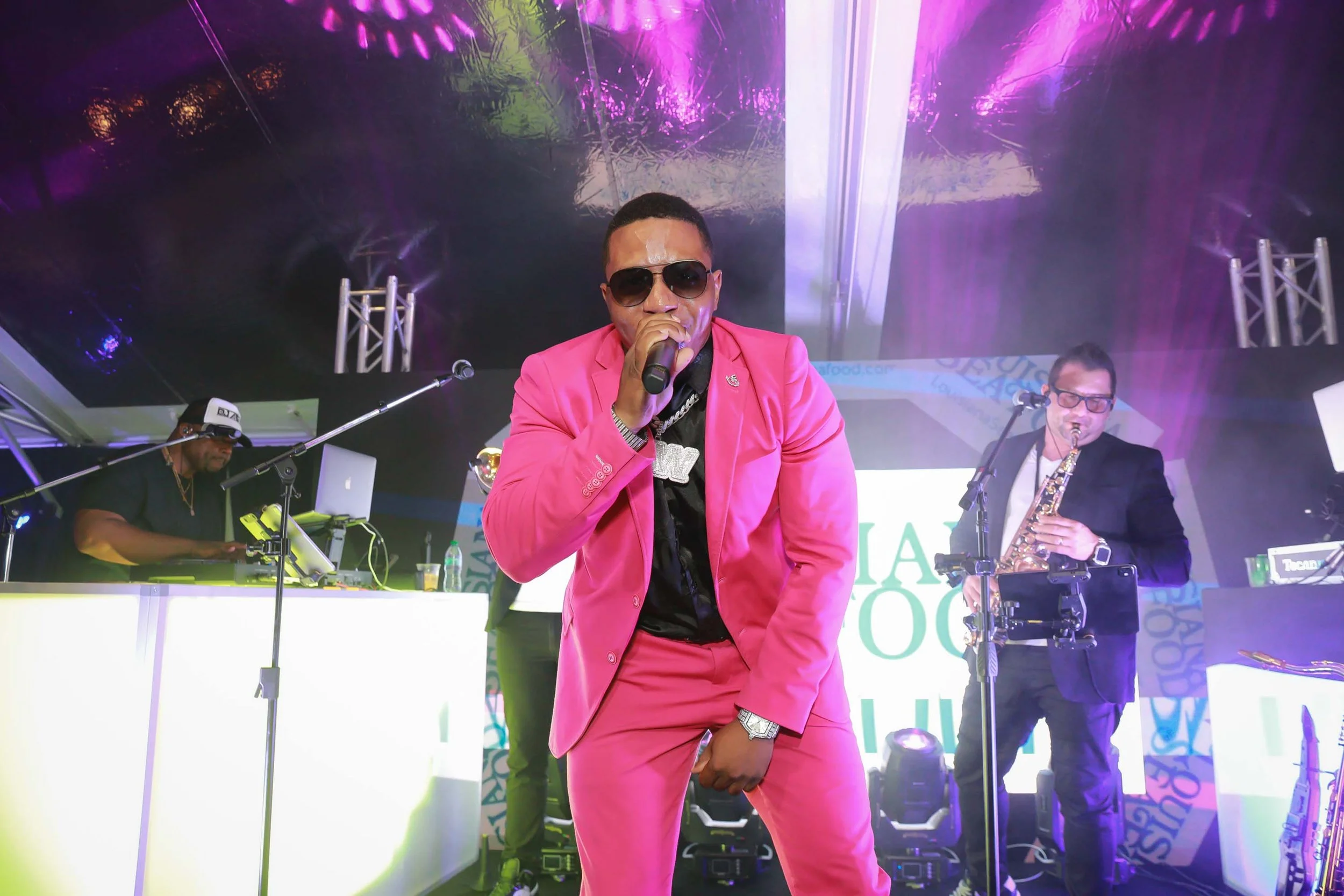 A male performer in a pink suit and sunglasses singing into a microphone during a live music performance with a band playing instruments behind him under purple lighting.