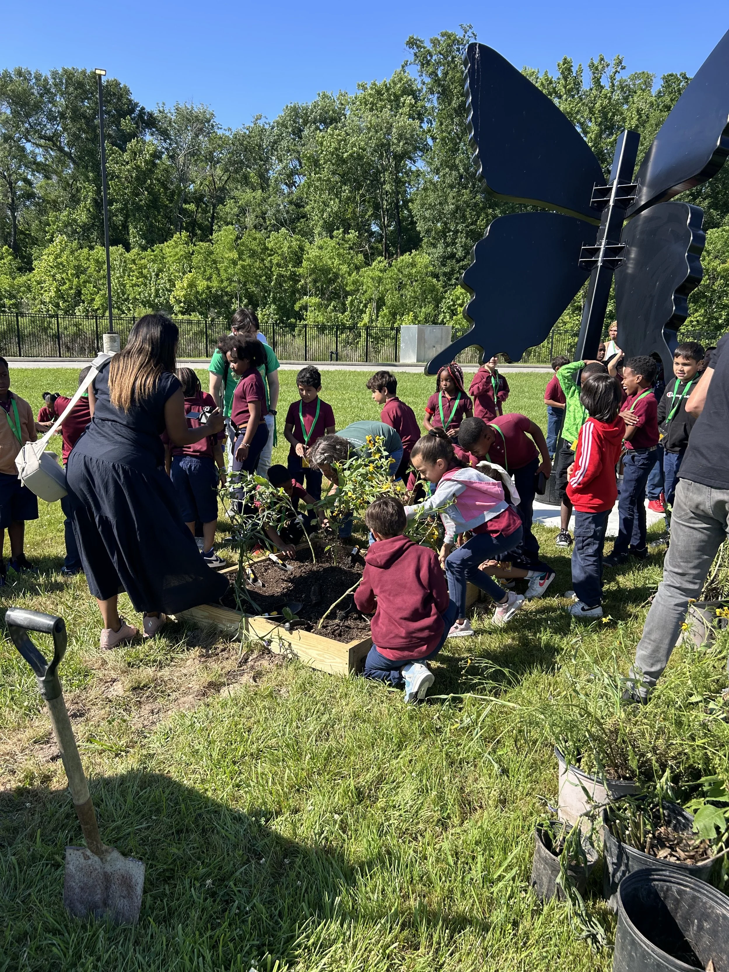 Group of children and adults planting a small tree in an outdoor garden on a sunny day, with large black butterfly sculpture and green trees in the background.