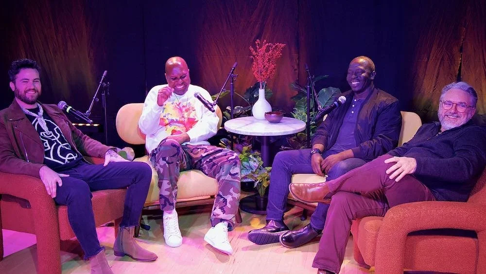 Four men sitting on chairs and sofa, smiling, with microphones in front of them, a small table with a vase and plants behind them, on a colorful stage.