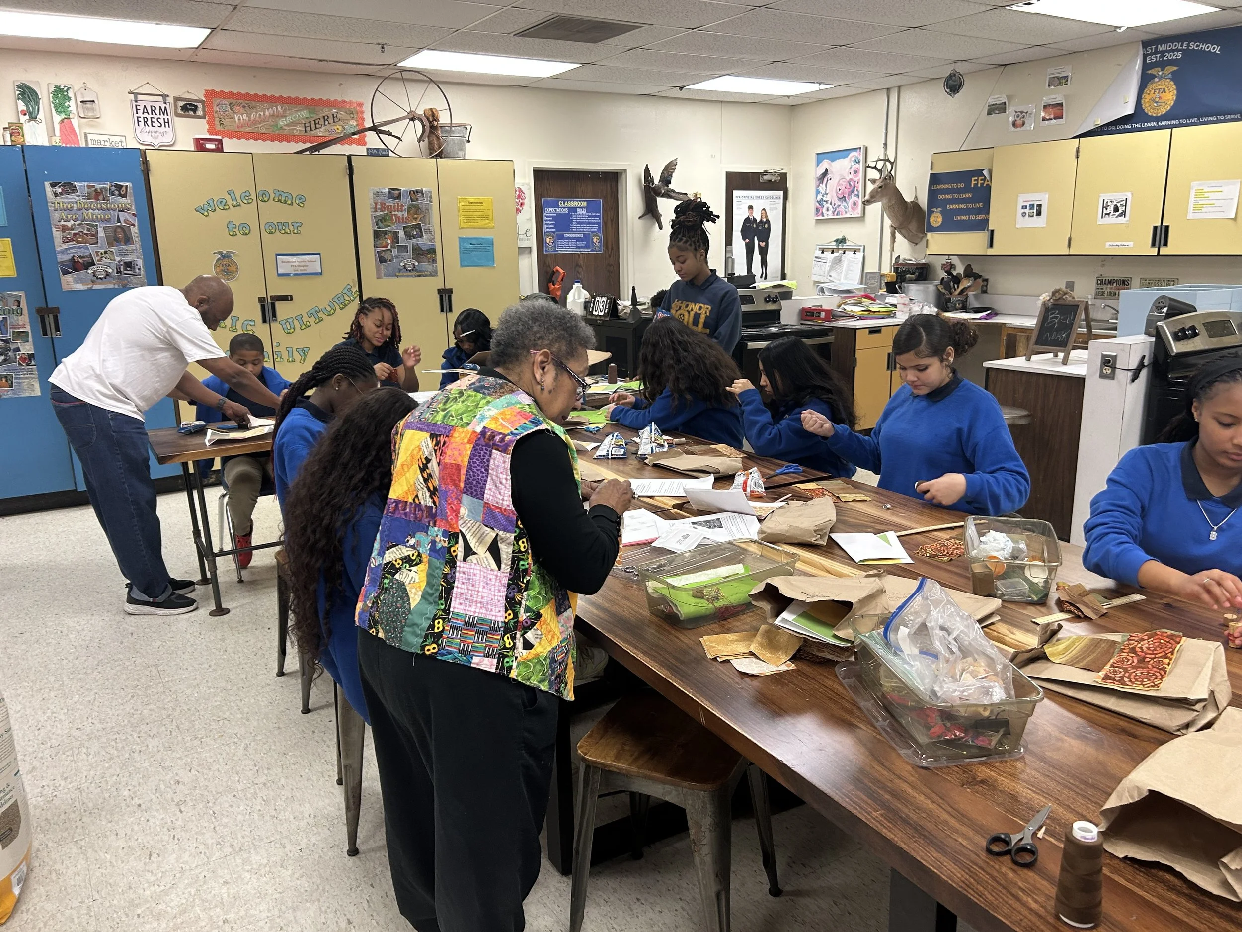 A group of students and adults participating in arts and crafts activities at a long table in a classroom, with various supplies and materials spread across the table.