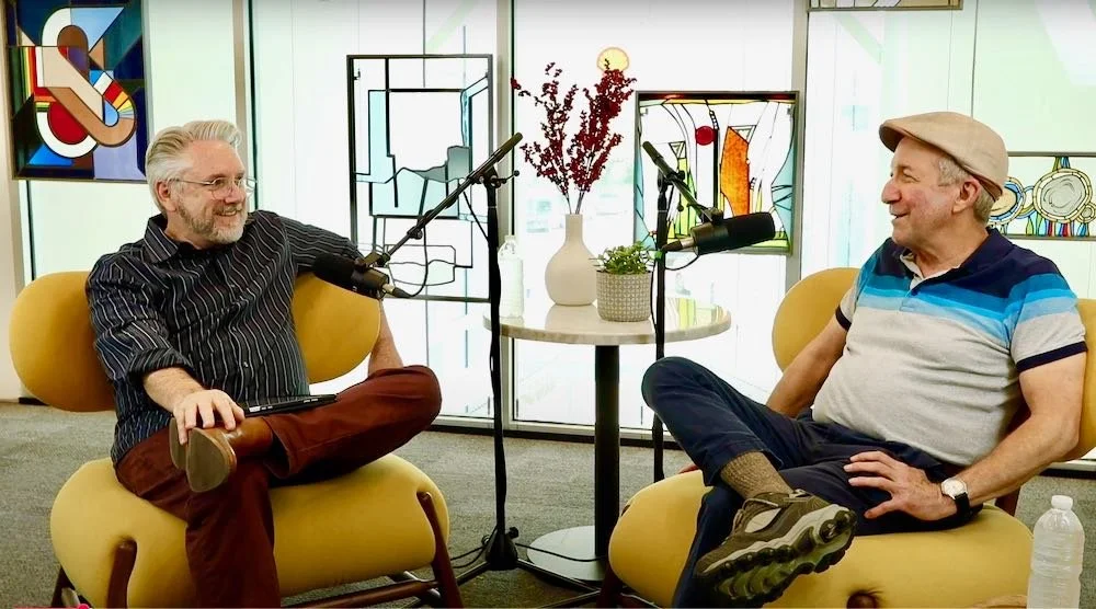 Two men sit on yellow chairs in a podcast studio, chatting and smiling. They are surrounded by microphones, a small table with a vase of flowers, and colorful stained-glass window art in the background.