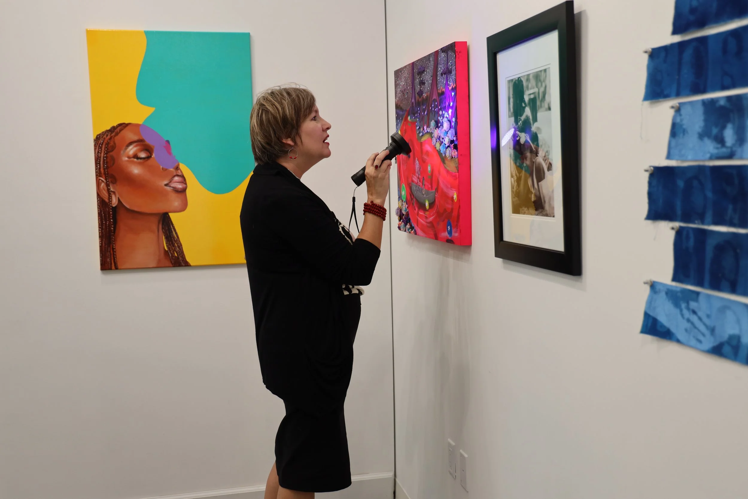 A woman with short hair and hoop earrings observes artwork on a gallery wall, holding a flashlight to illuminate the pieces.