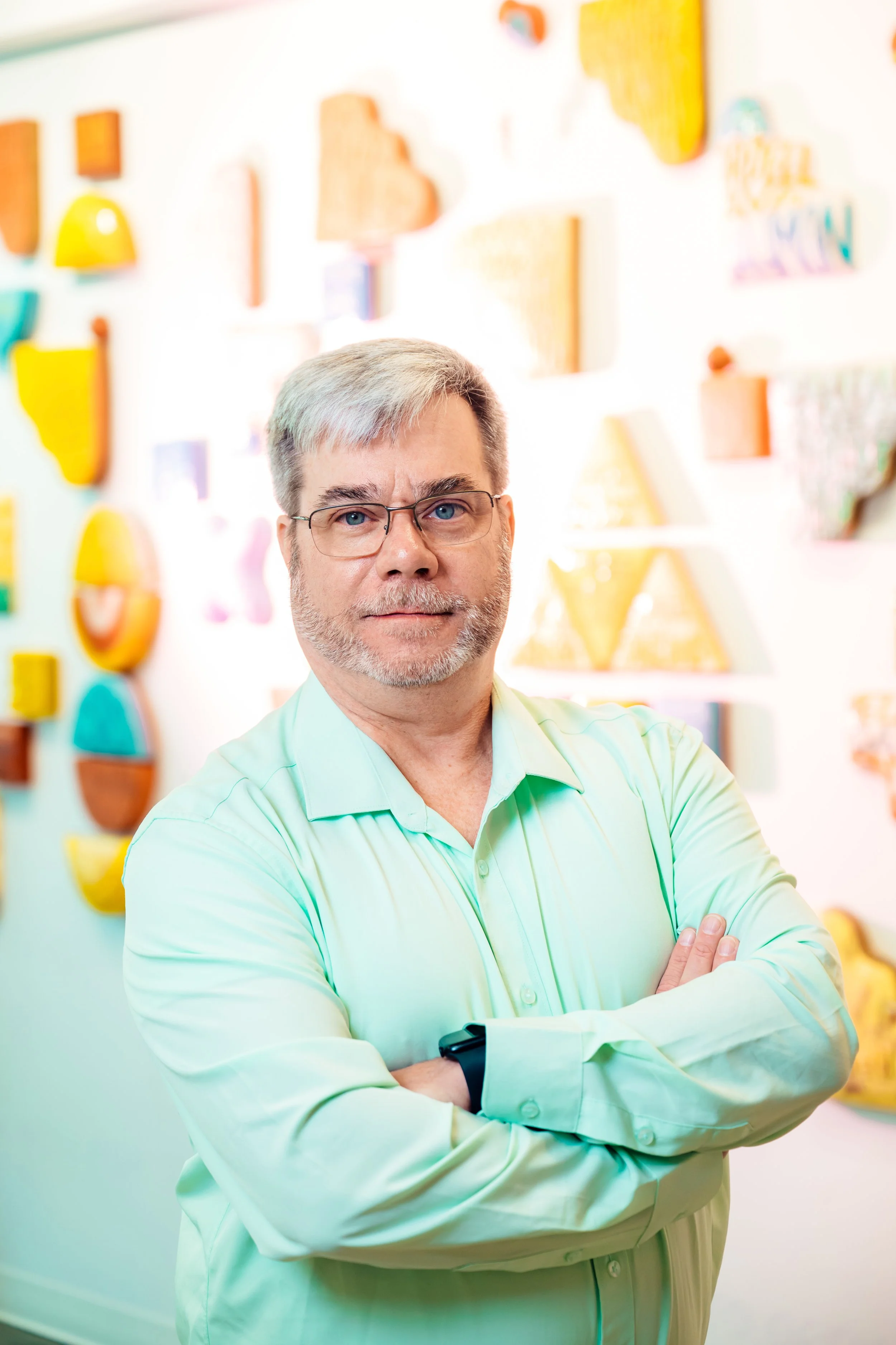 A man with glasses and gray hair, wearing a light green shirt, standing with arms crossed in front of a colorful abstract wall art display.