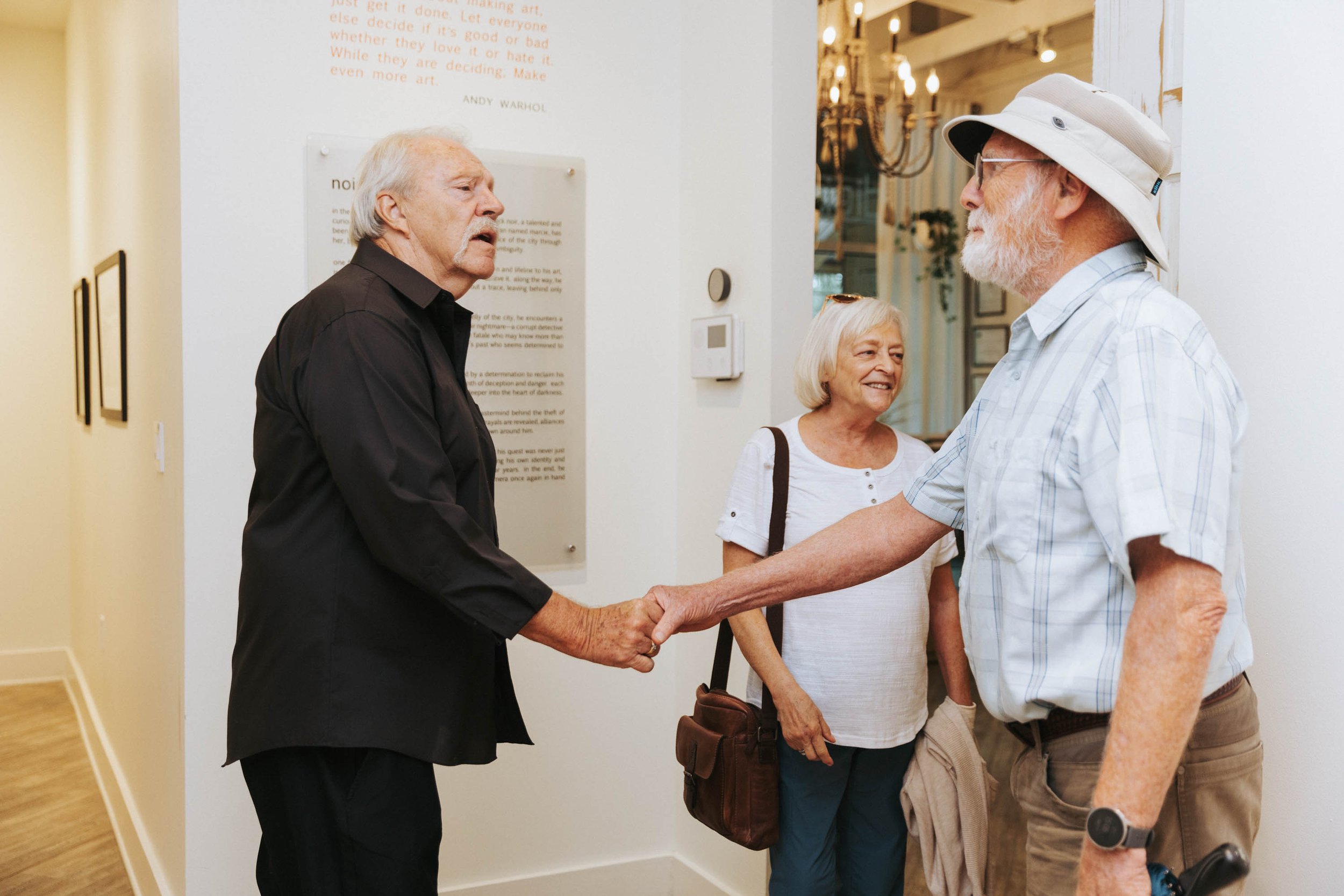 Older man in a black shirt shaking hands with another man wearing a white hat and glasses inside an art gallery, with a woman smiling behind them and text on the wall in the background.