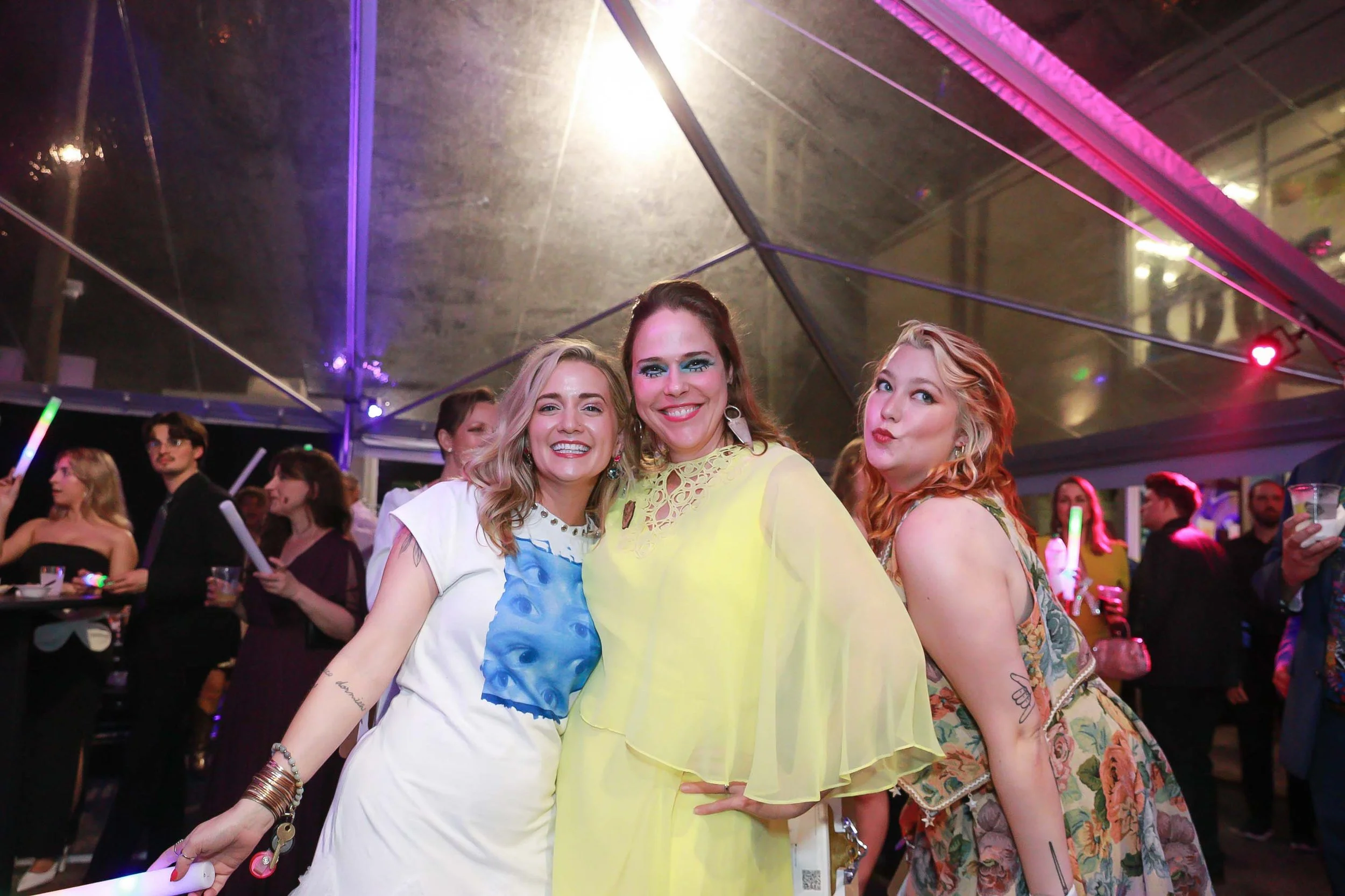 Three smiling women posing together at a party with colorful lights and other guests in the background.