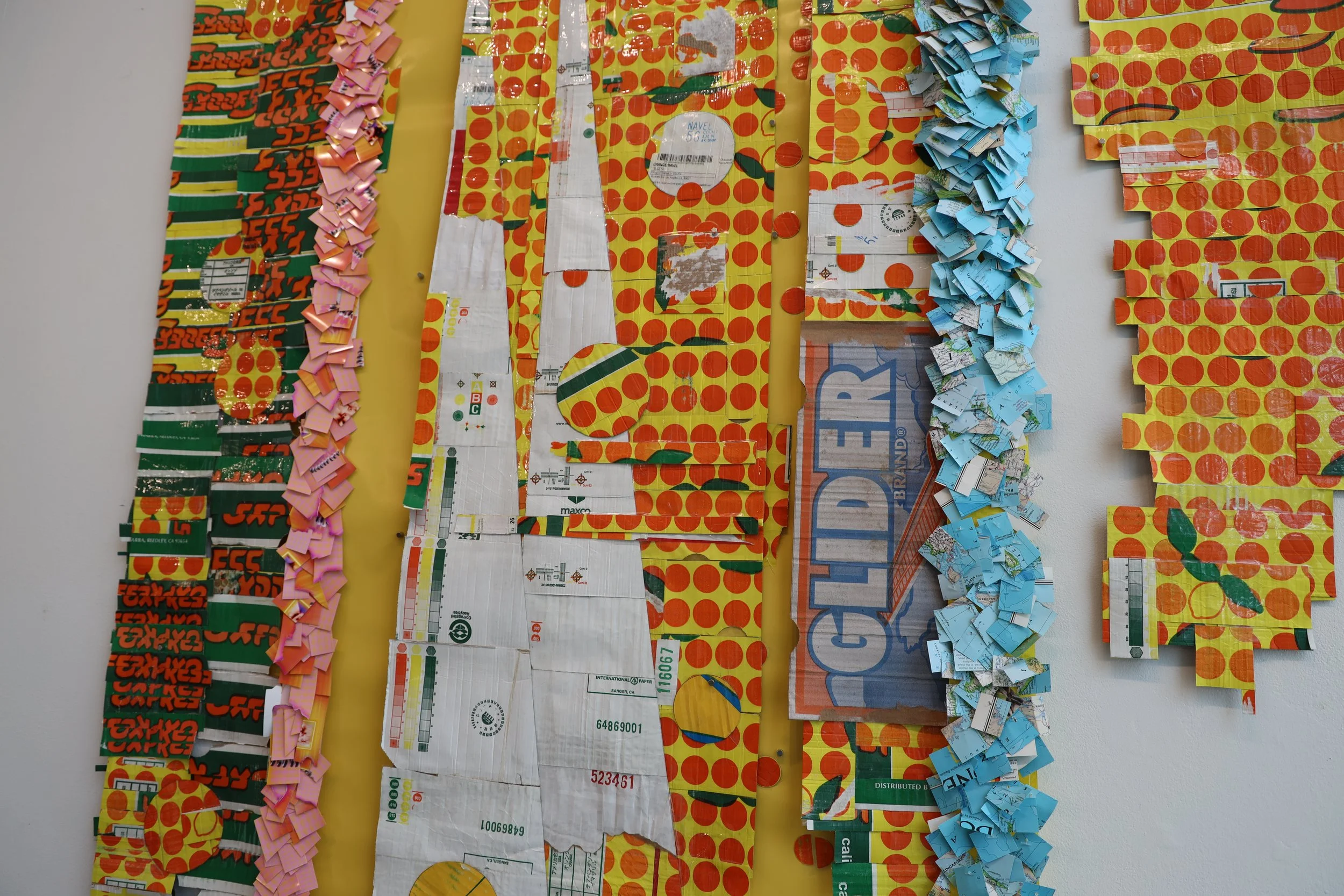 Multiple strips of original packaging, including flattened cardboard, paper, and plastic labels, arranged on a wall to create a collage. The packaging features bright colors such as yellow, red, green, and blue.