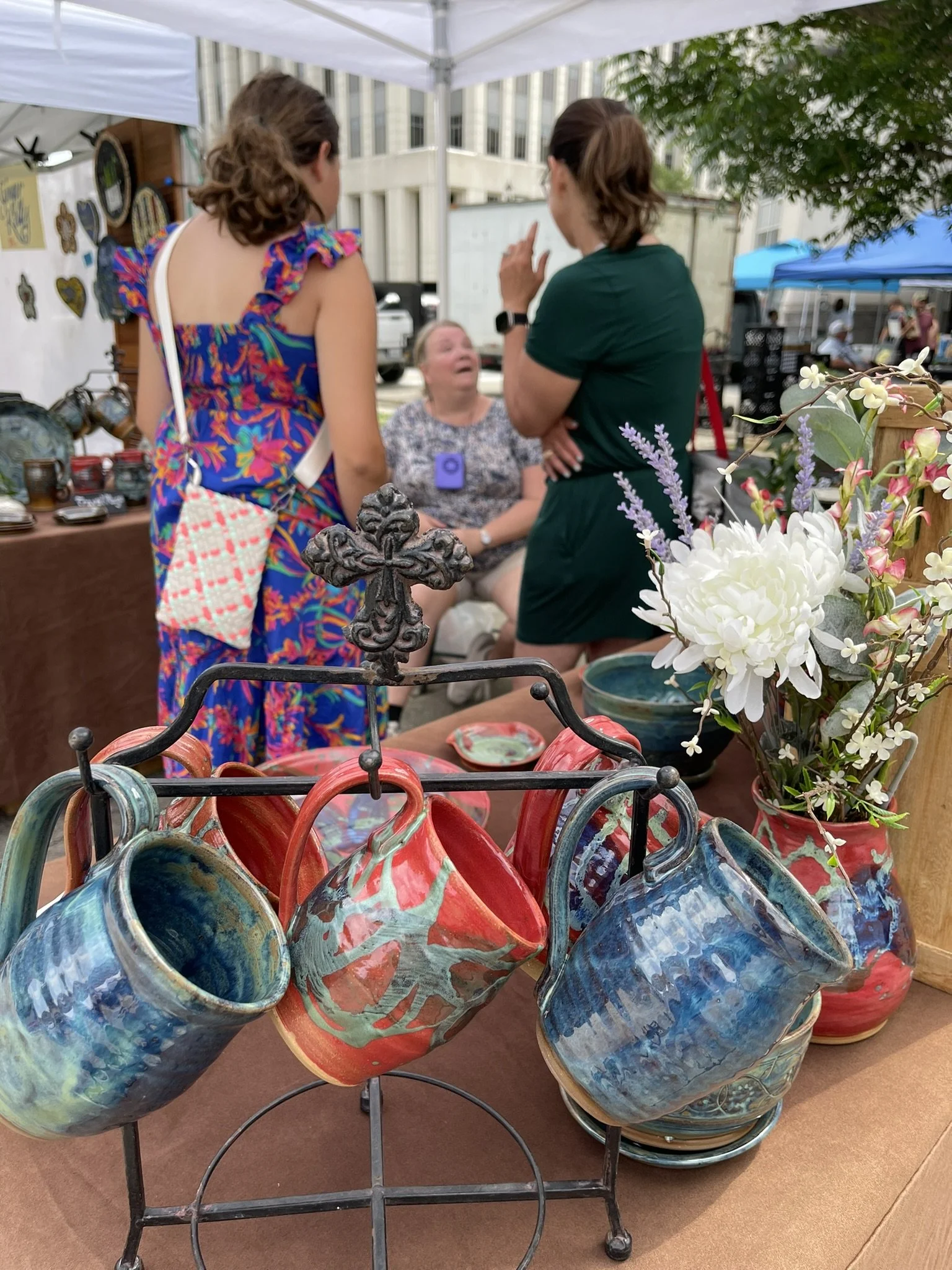 Colorful ceramic vases displayed at an outdoor market stall, with a background of people and tents, and a bouquet of flowers in the foreground.