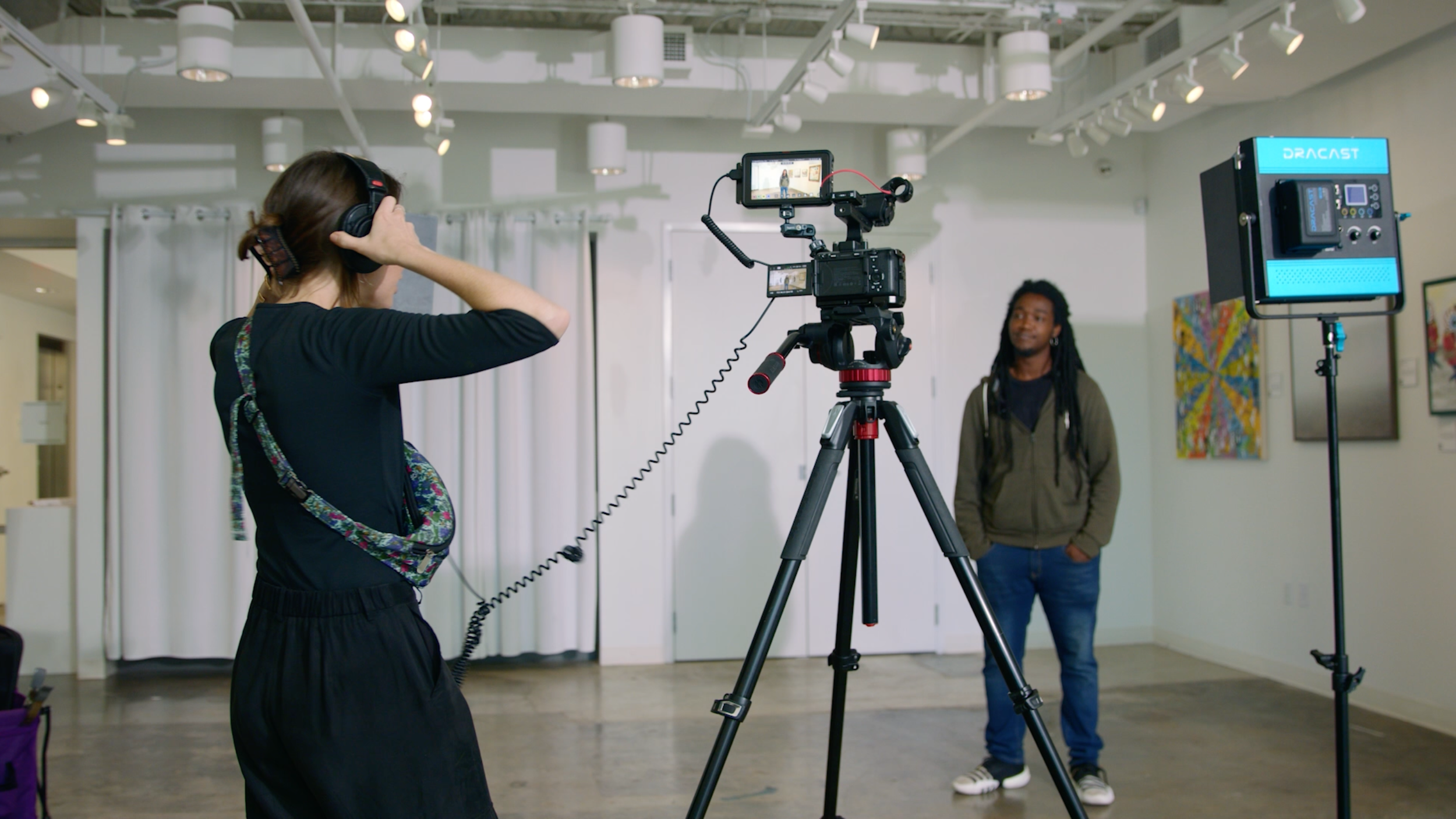 A woman wearing headphones adjusts a camera on a tripod as a man with dreadlocks watches in an art gallery or studio.