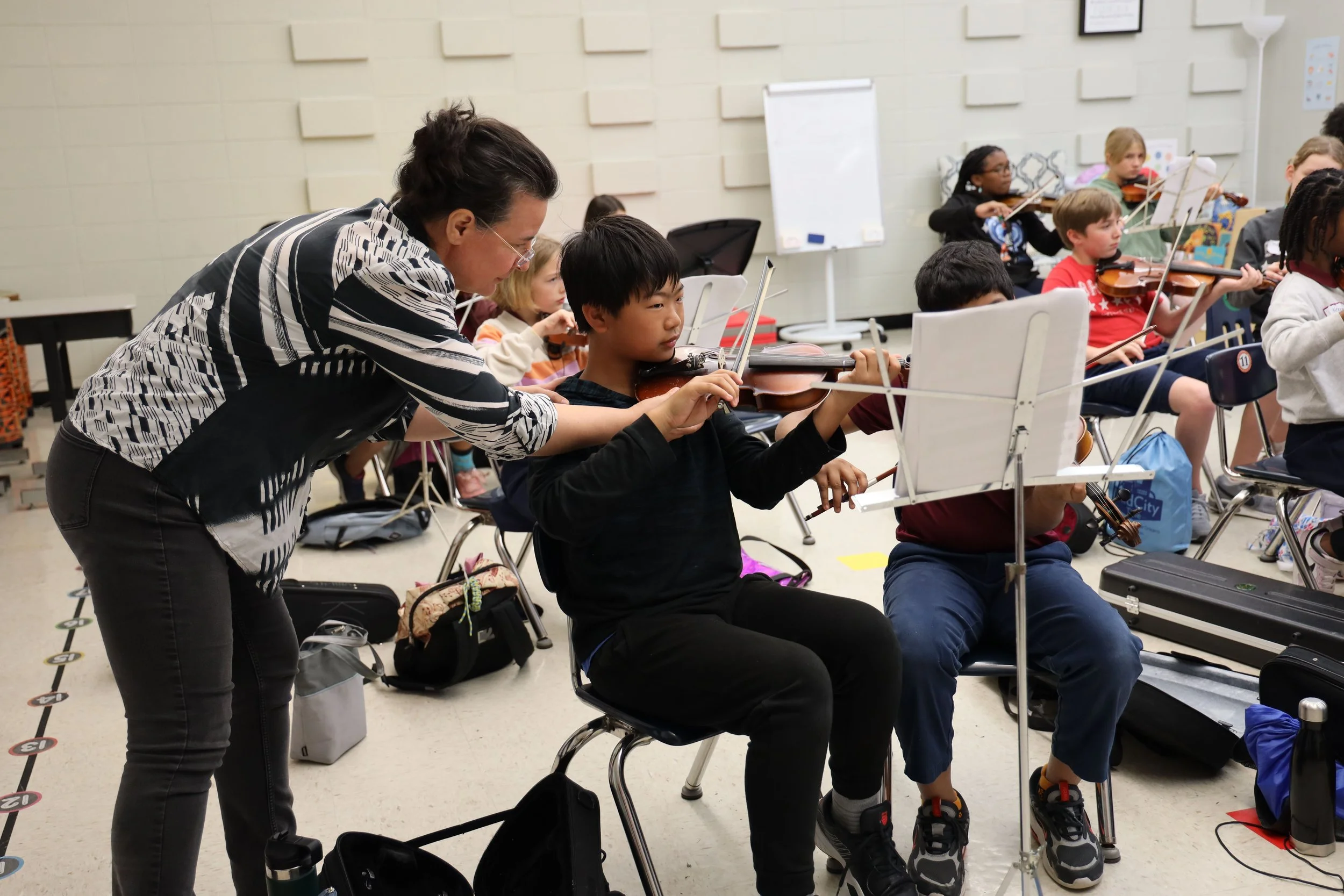 A music teacher helps a young boy with his violin in a classroom full of children playing violins.