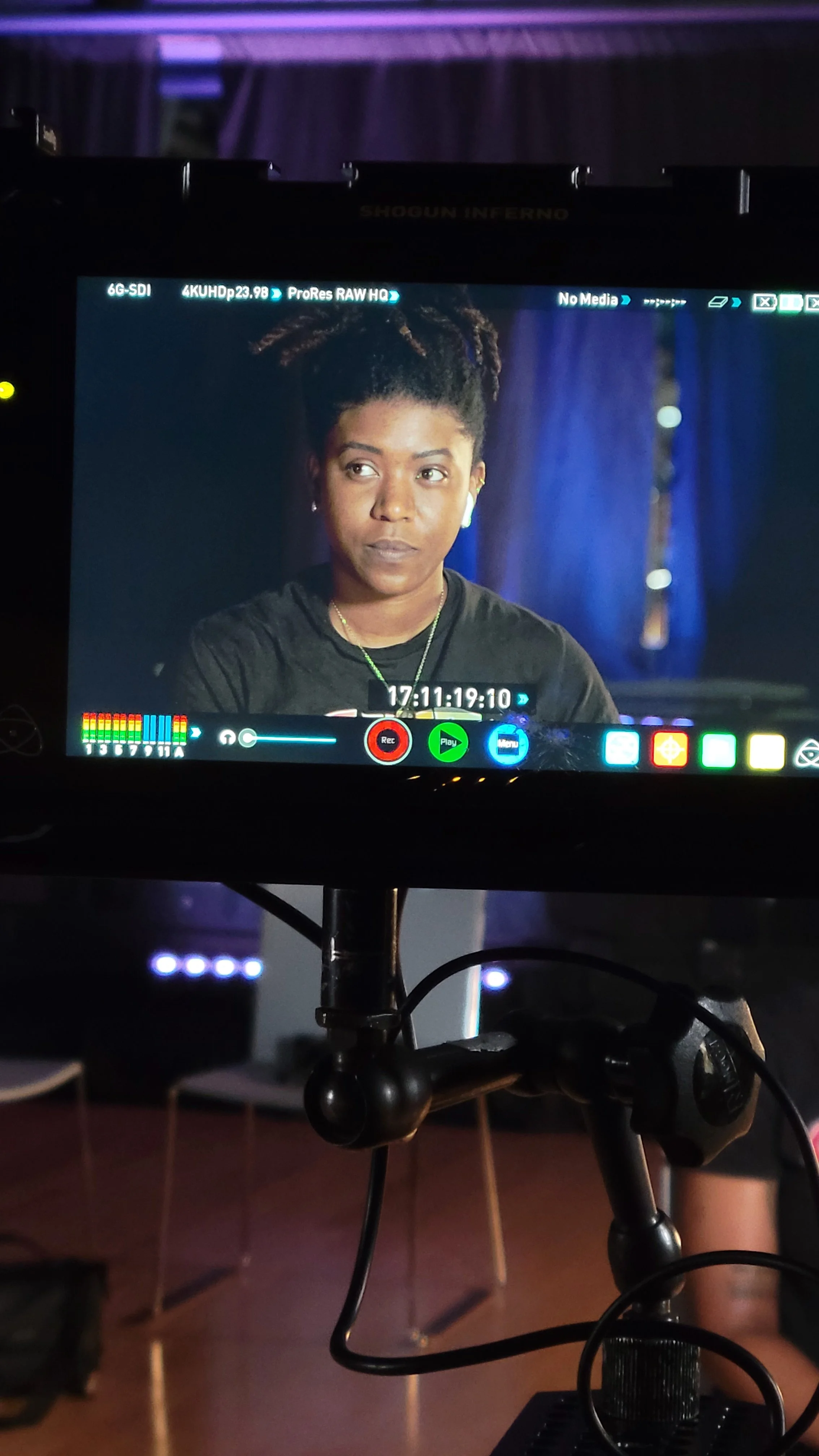 Camera monitor showing a woman with dark skin and natural hair styled in short twists, wearing a black shirt and gold jewelry, with a blue curtain in the background.